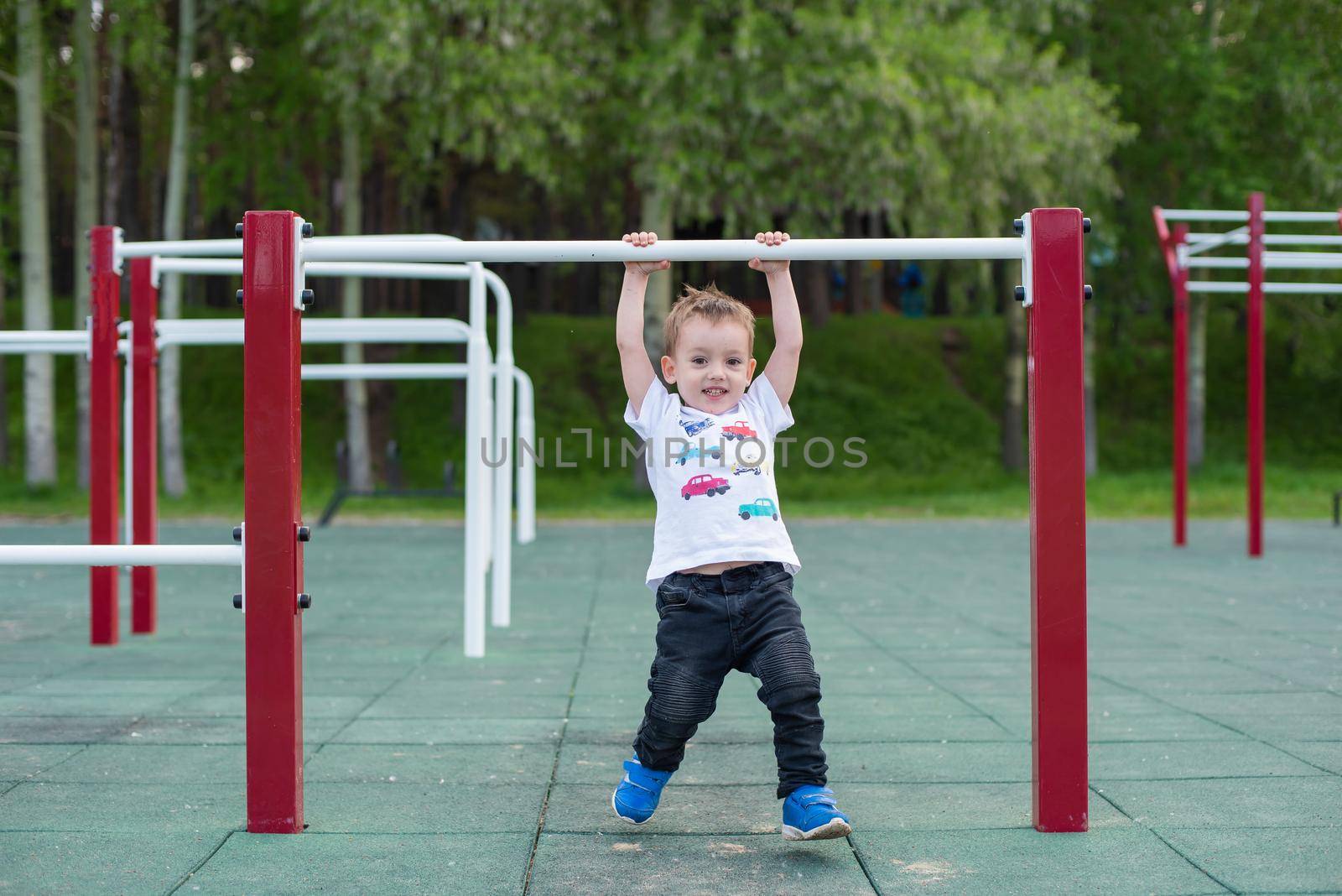 A little boy learns to pull up on a horizontal bar in the open air. The child is hanging on the uneven bars