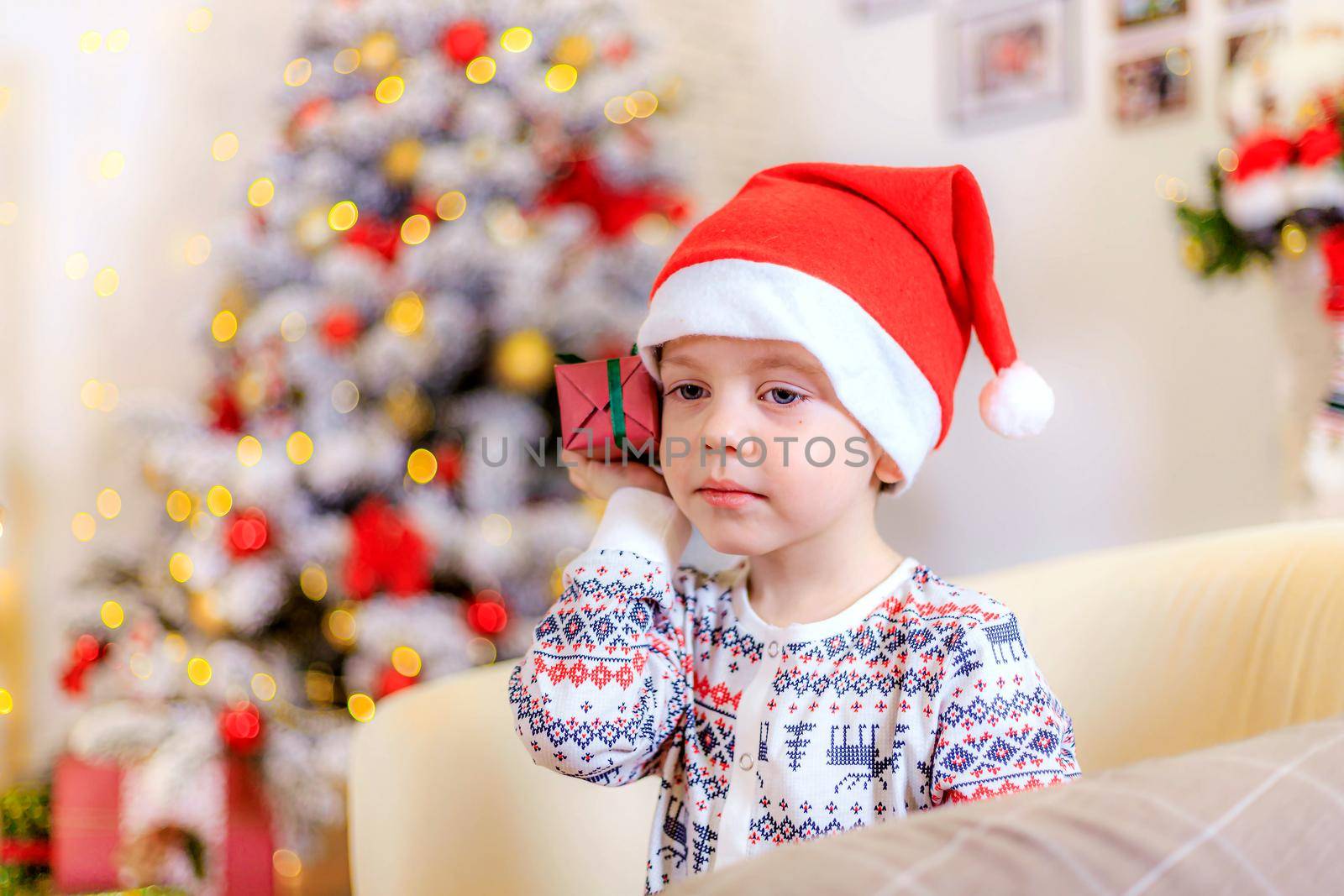 Boy at the Christmas tree. The boy folded his hands.. Cute baby . Holiday. Article about new year and Christmas. Colorful spruce. Garland. Holiday decoration and decor . Santa hat.
