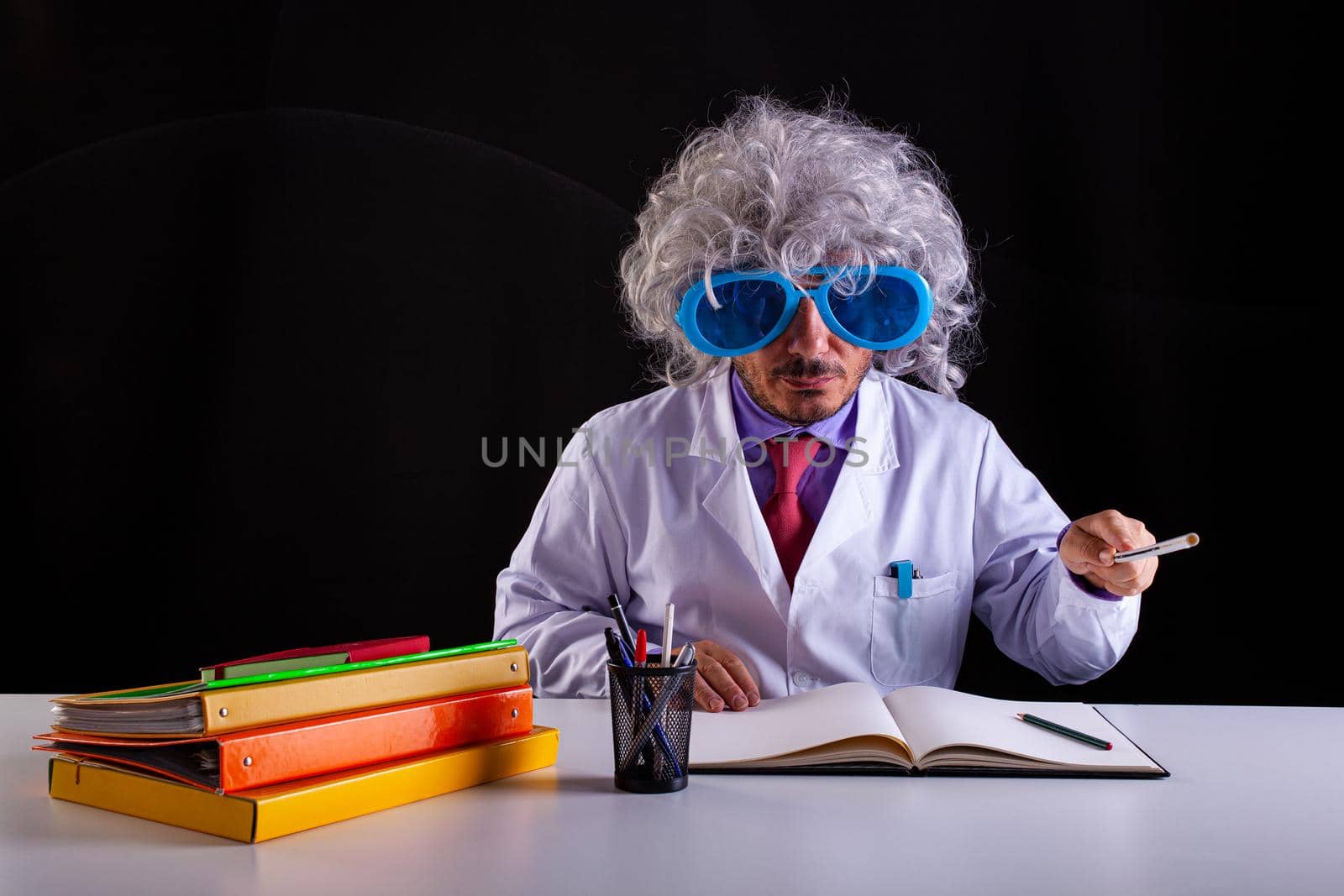 Crazy science teacher in white coat with unkempt hair in funny eye glasses sitting at the desk