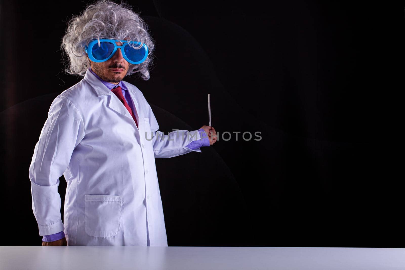 Crazy science teacher in white coat with unkempt hair in funny eye glasses holding a wand to point at the blackboard