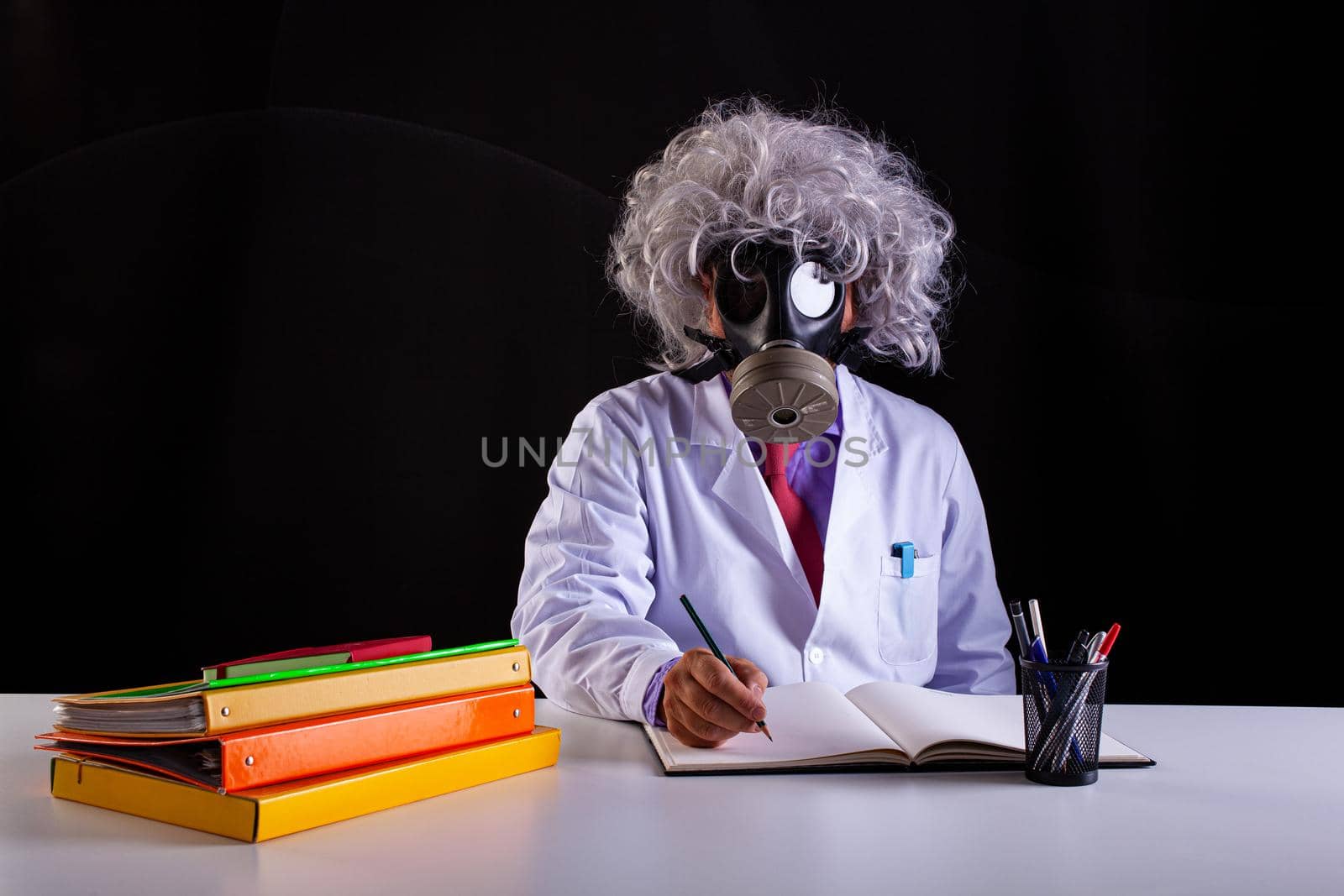 Crazy science teacher in white coat with unkempt hair sitting at the desk wears a gas mask by bepsimage