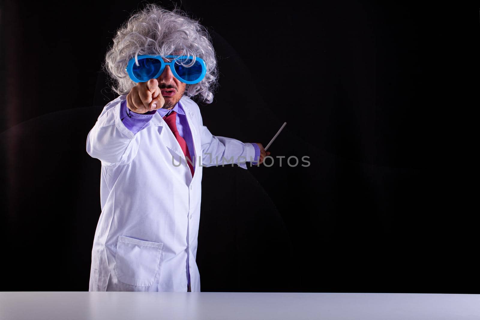 Angry science teacher in white coat with unkempt hair in funny eye glasses points with his finger on black background