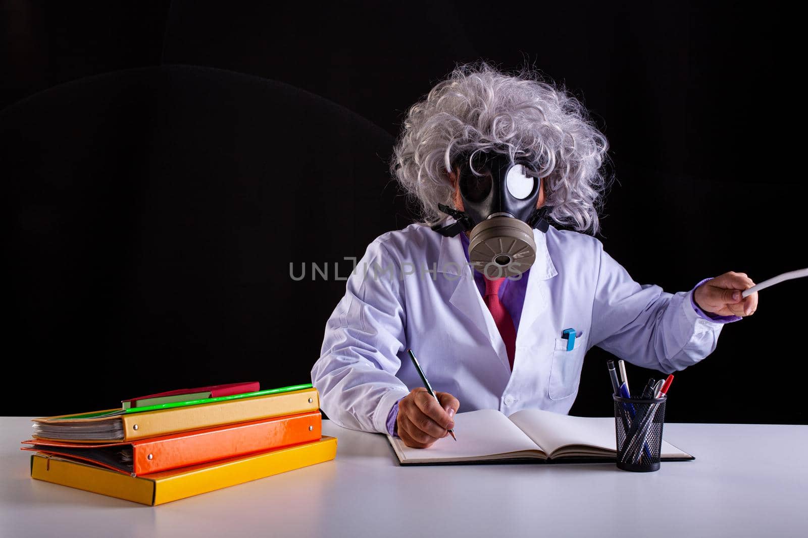 Crazy science teacher in white coat with unkempt hair sitting at the desk wears a gas mask