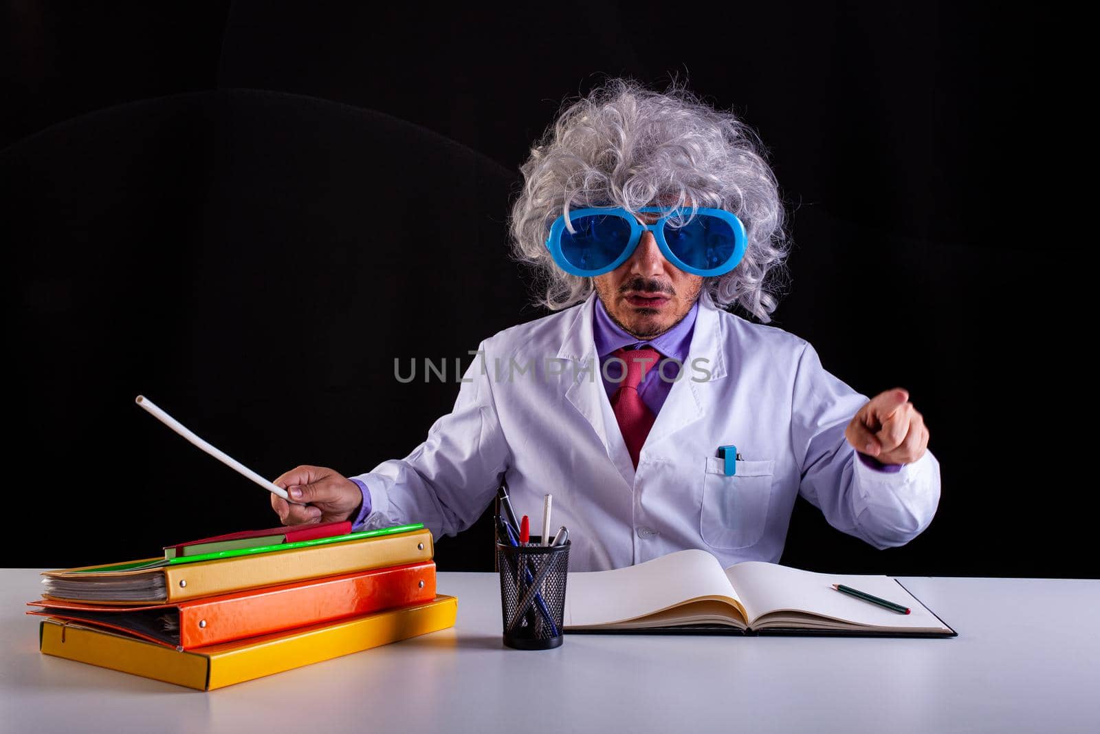 Crazy science teacher in white coat with unkempt hair in funny eye glasses sitting at the desk