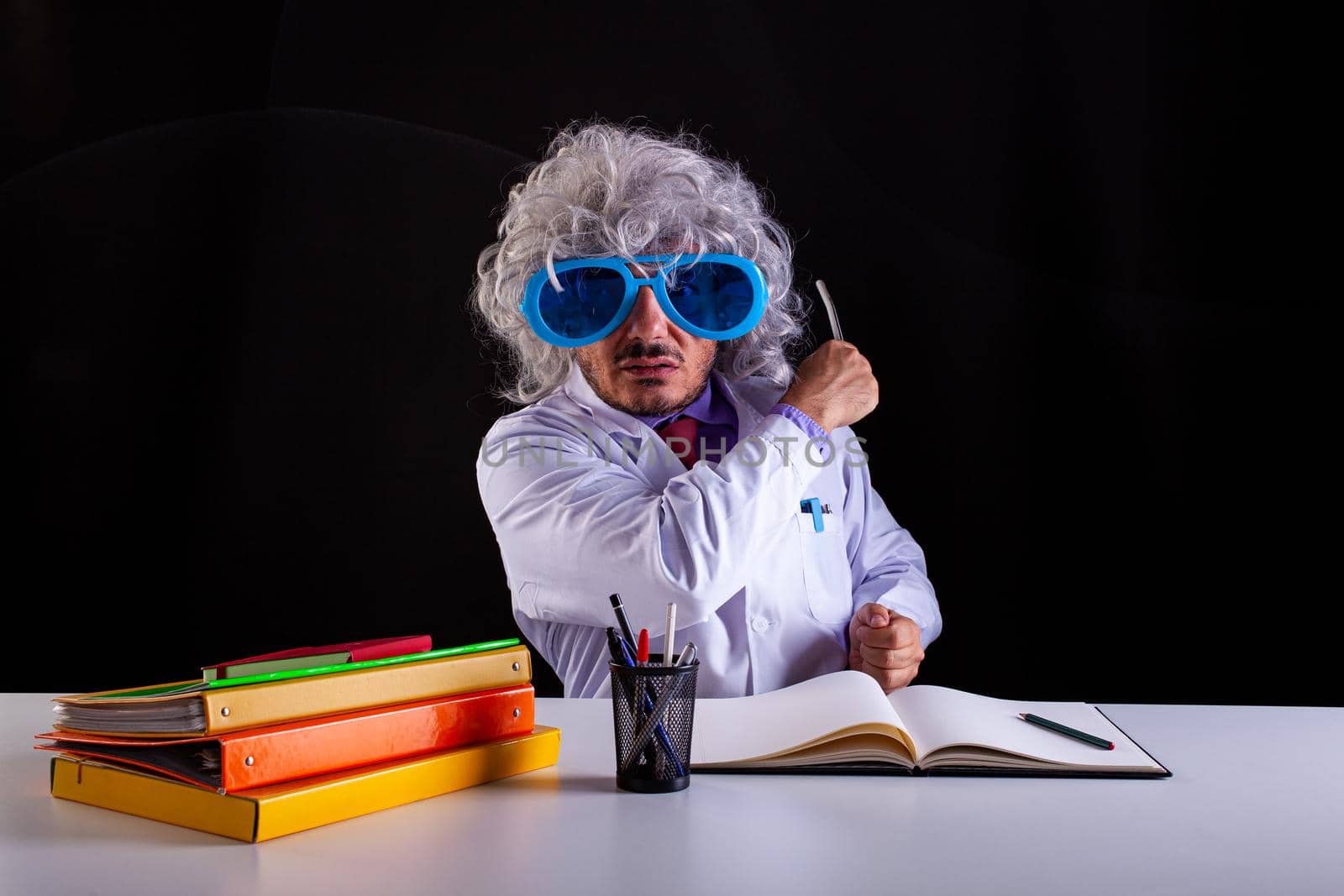 Crazy science teacher in white coat with unkempt hair in funny eye glasses sitting at the desk holding a wand to point at the blackboard