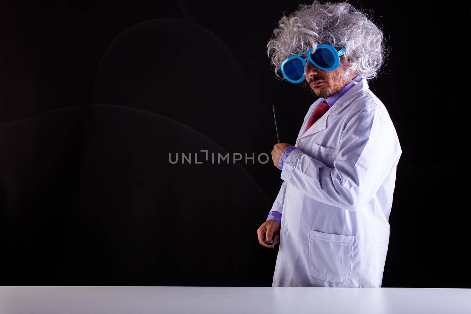 Crazy science teacher in white coat with unkempt hair in funny eye glasses holding a wand to point at the blackboard