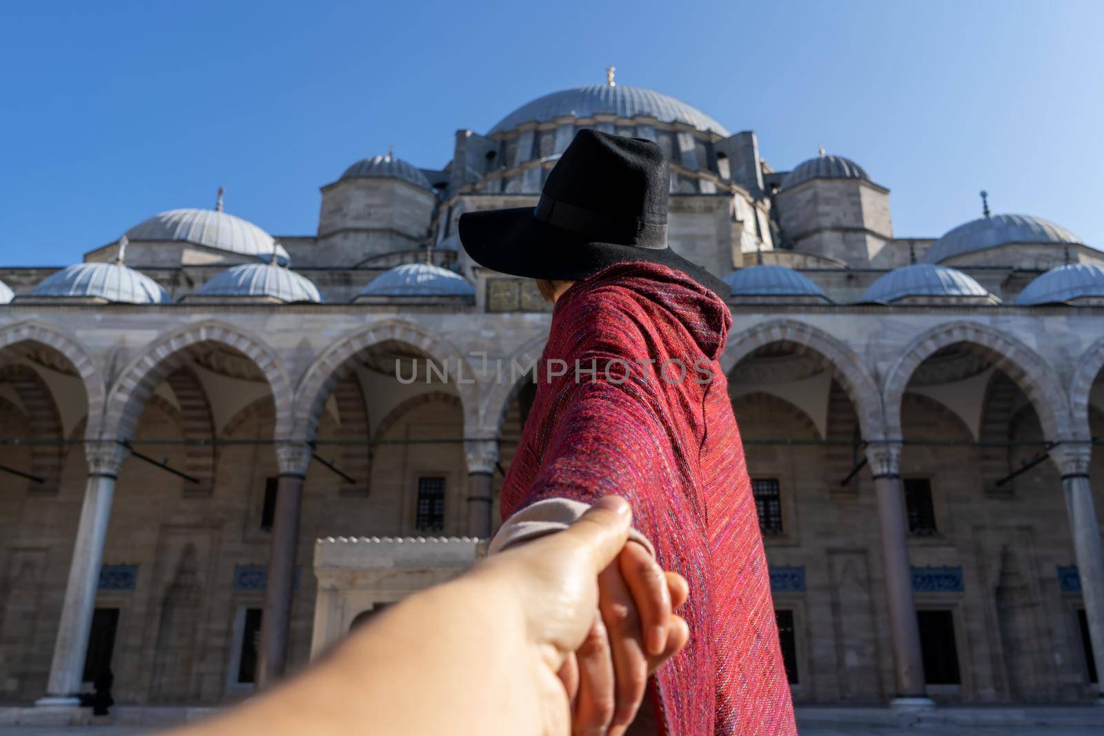 A young traveler girl in a red poncho and hat holds a friend's hand, follow me gesture. A couple in love travels to Arab countries. An ancient blue mosque on the background, Istanbul, Turkey.