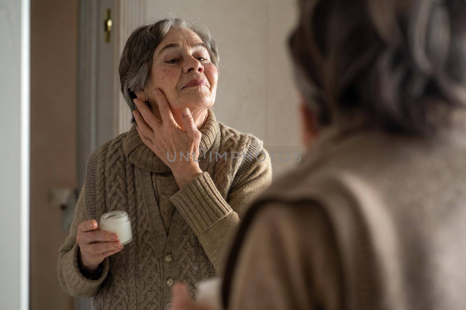 An elderly woman applies an anti-aging moisturizer. The pensioner looks after her appearance, makes cosmetic procedures in the bathroom. Uses cream for skin rejuvenation, regeneration, lifting.