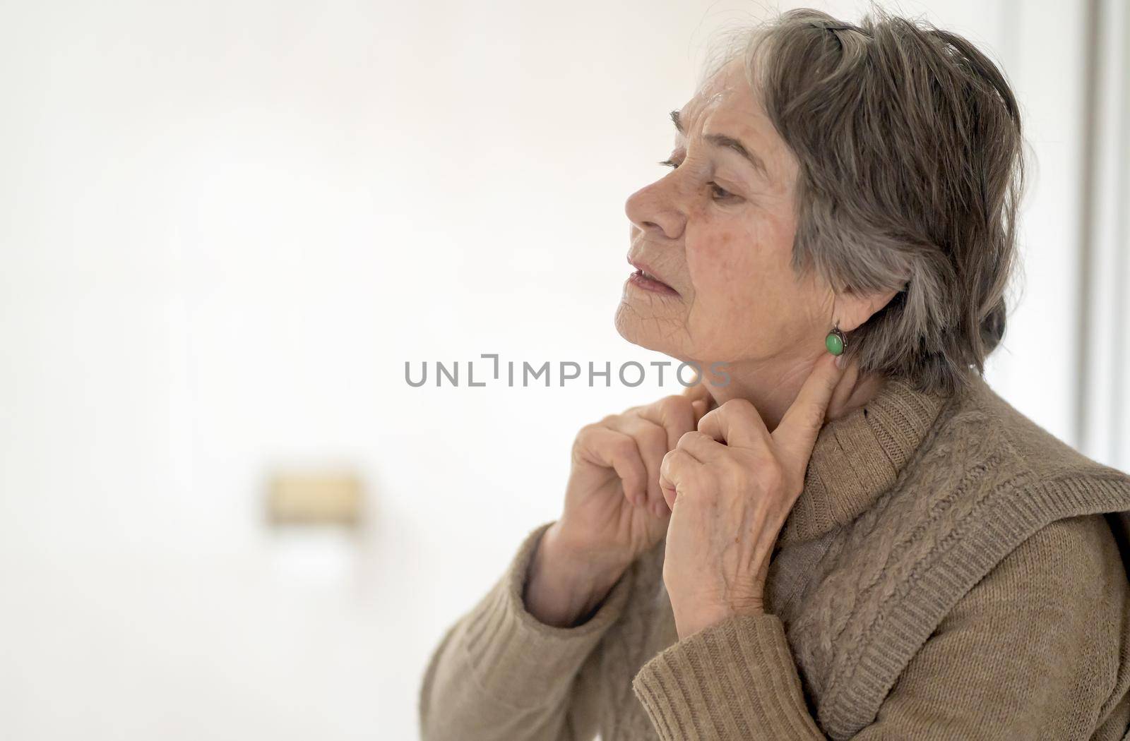 An elderly woman applies an anti-aging An elderly woman puts fingers on the lymph nodes and looks at the mirror. Takes care, monitors thyroid hormones and the health of the endocrine system.