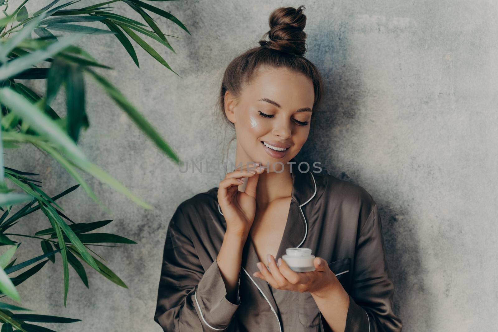 Beautiful europian brunette woman at home in silk brown pajama doing beauty procedures, applying moisturizing face cream during morning make up routine. Skincare concept