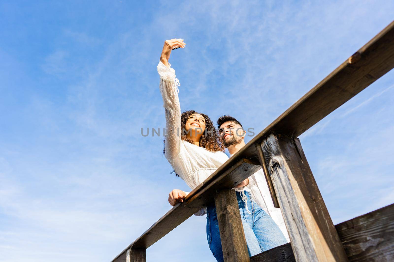 Unusual low angle view shot of multiracial couple on a wooden beach bridge taking a selfie in vacation at sea ocean resort. Large sky copy space ideal for banner or ads. Hispanic girl using smartphone