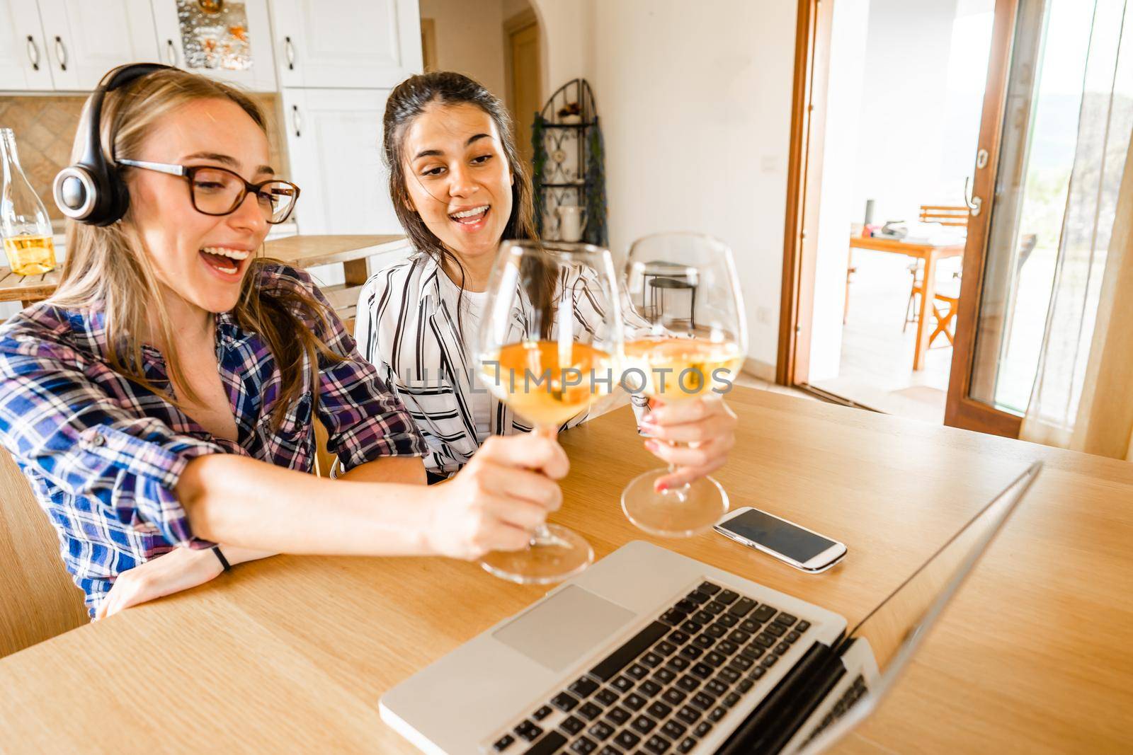 Two happy young women at home sitting at table looking laptop toasting with champagne or white wine glasses. New normal celebration online due to social distancing and video conferencing technology by robbyfontanesi