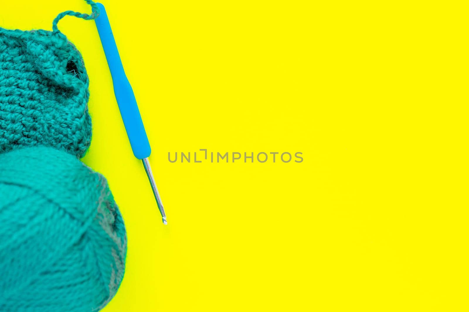 A metal hook with a blue handle, a skein of green yarn and a part of a knitted fragment lies on the side of the photo on a yellow background. top view, flat lay, copy space, isolate. High quality photo