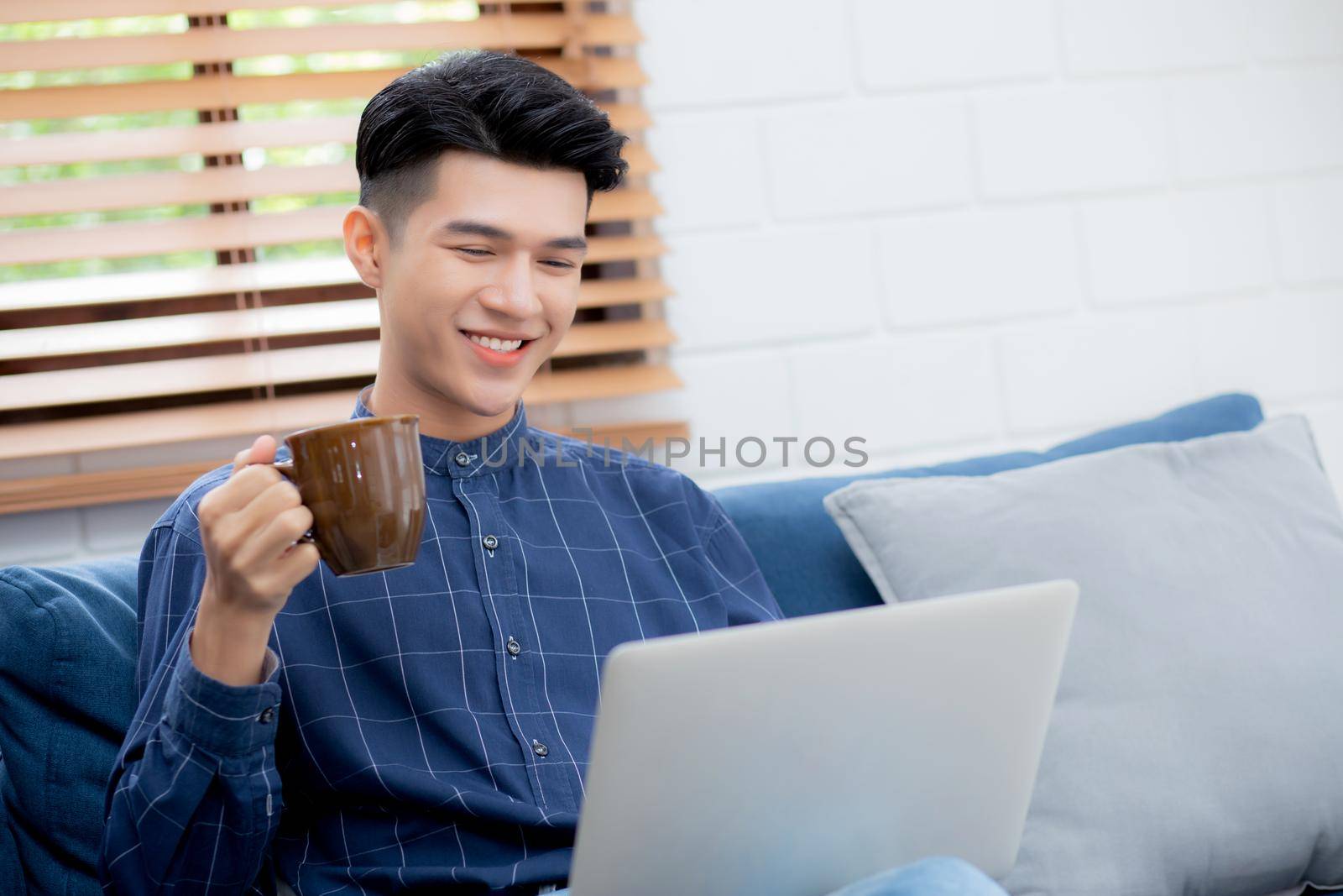 Young asian businessman smile and work from home with laptop computer online on sofa in living room, freelance business man using notebook and drink coffee on couch, new normal, lifestyle concept. by nnudoo