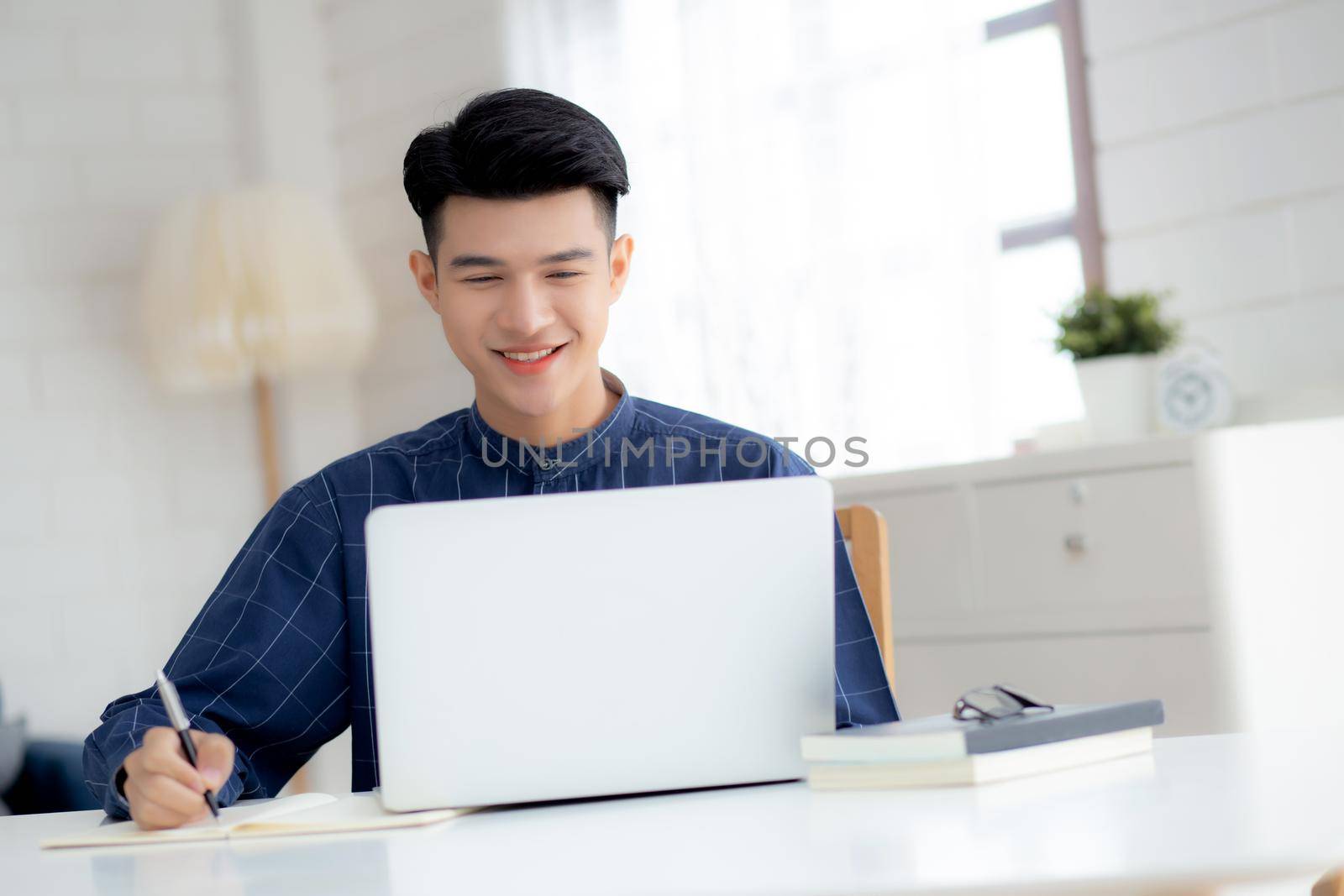 Young asian business man writing on notebook for planning working and using laptop computer on desk at home, notes about finance, male study and learning, business and communication concept.