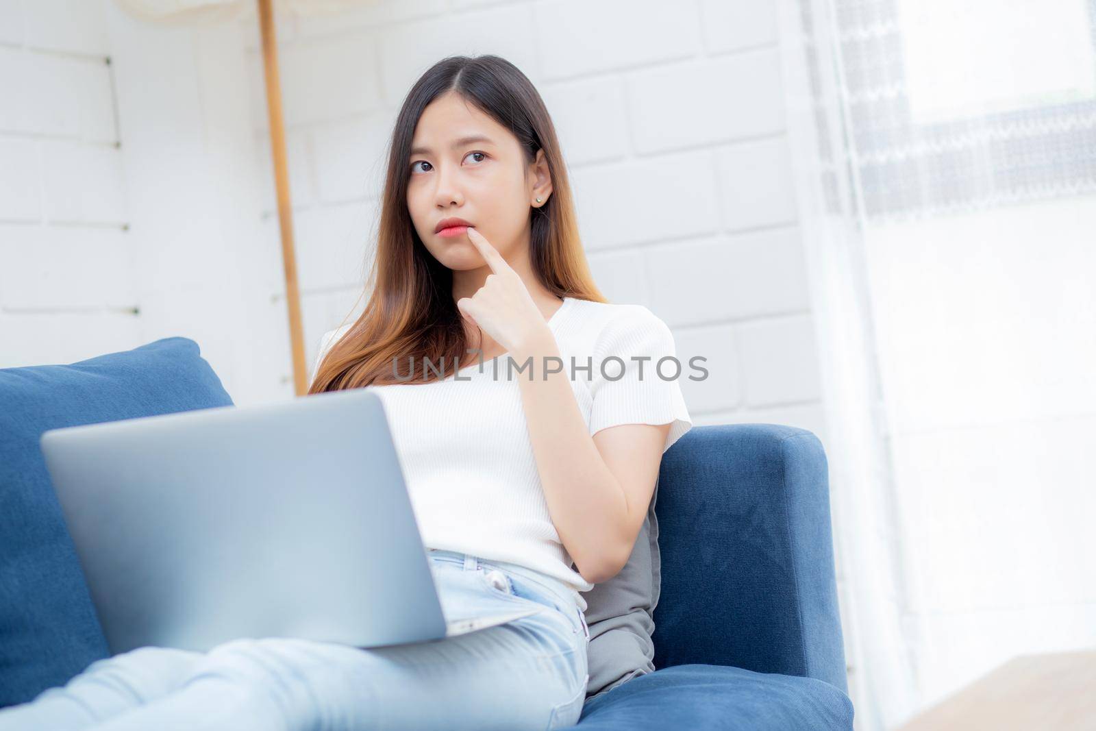 Beautiful young asian woman working on laptop computer and thinking idea on sofa at home, freelance girl sitting on couch using notebook to internet at living room, one person, lifestyle concept.