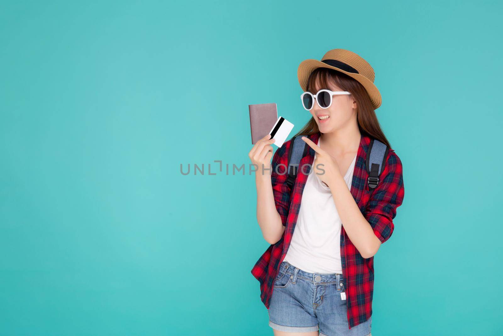 Beautiful young asian woman wear hat and sunglasses with showing credit card and passport and pointing to travel summer trip isolated on blue background, girl in vacation with payment finance on card. by nnudoo