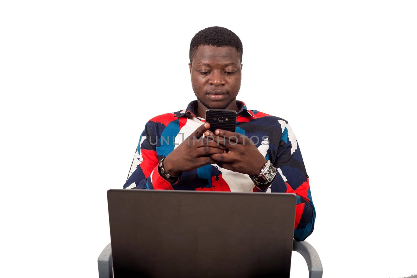 young african sitting in multicolored shirt with laptop on thighs watching mobile phone.