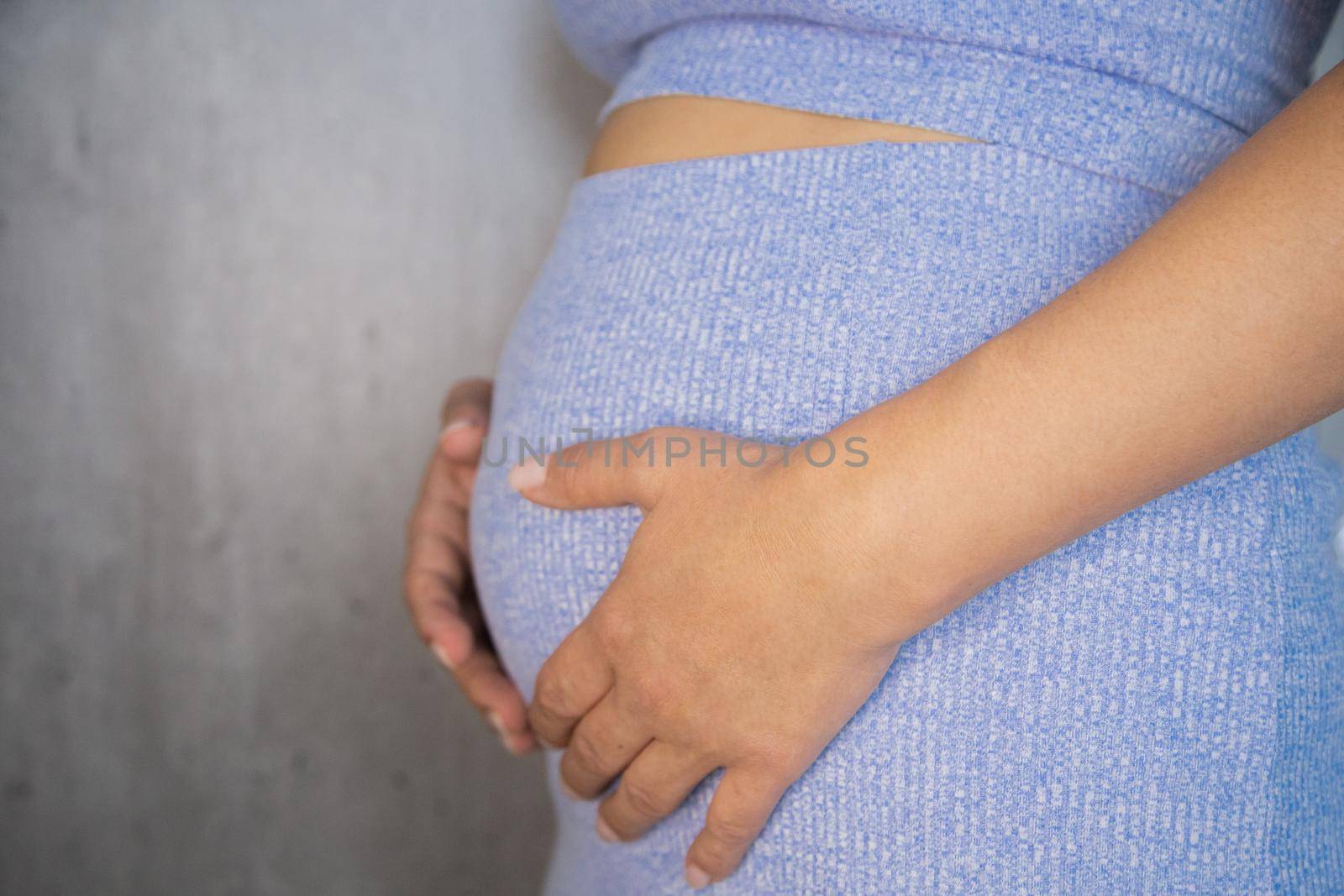 A pregnant woman is standing in a blue suit and holding her tummy with her hands on a gray background. by Kseniia12393