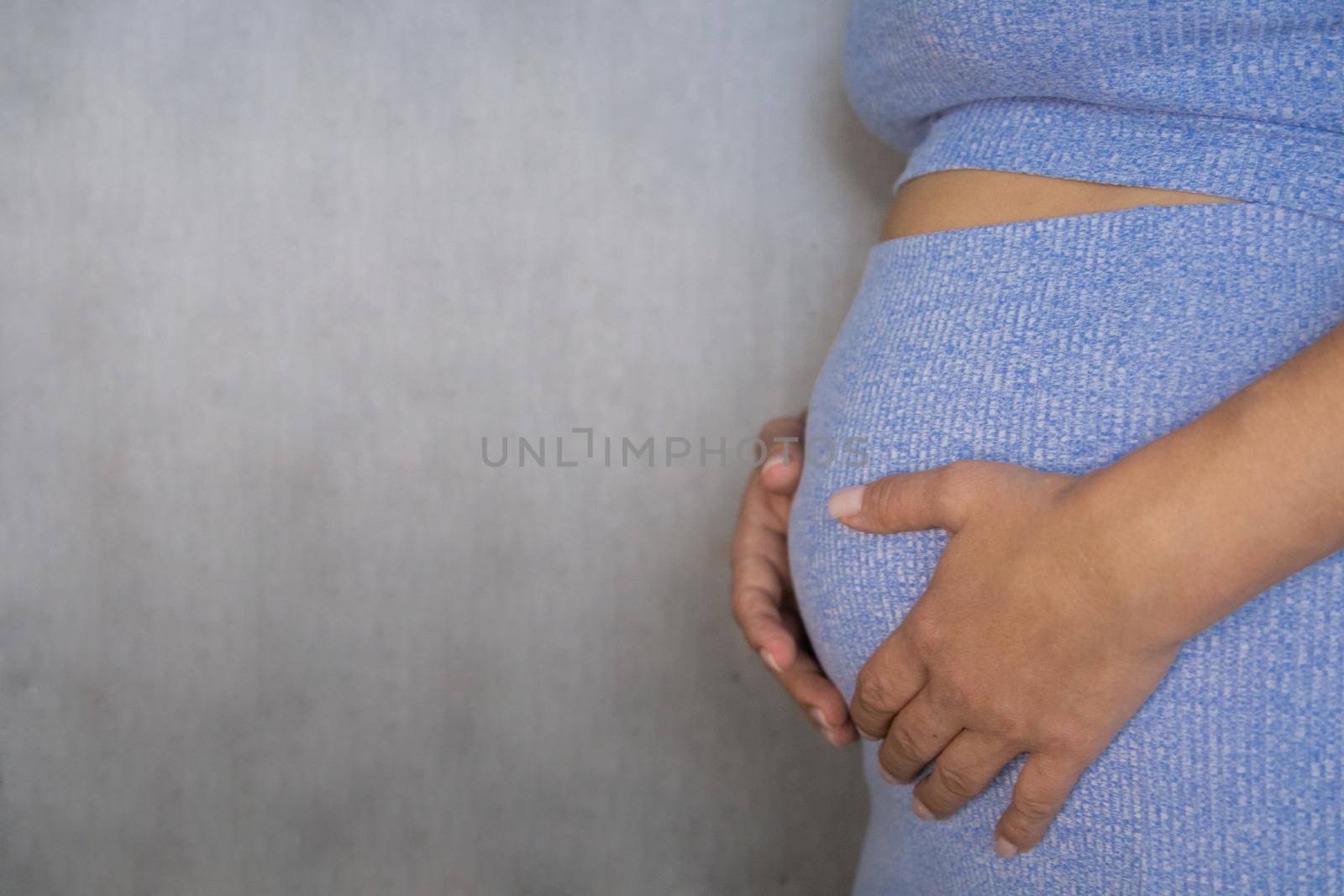 A pregnant woman is standing in a blue suit and holding her tummy with her hands on a gray background. by Kseniia12393