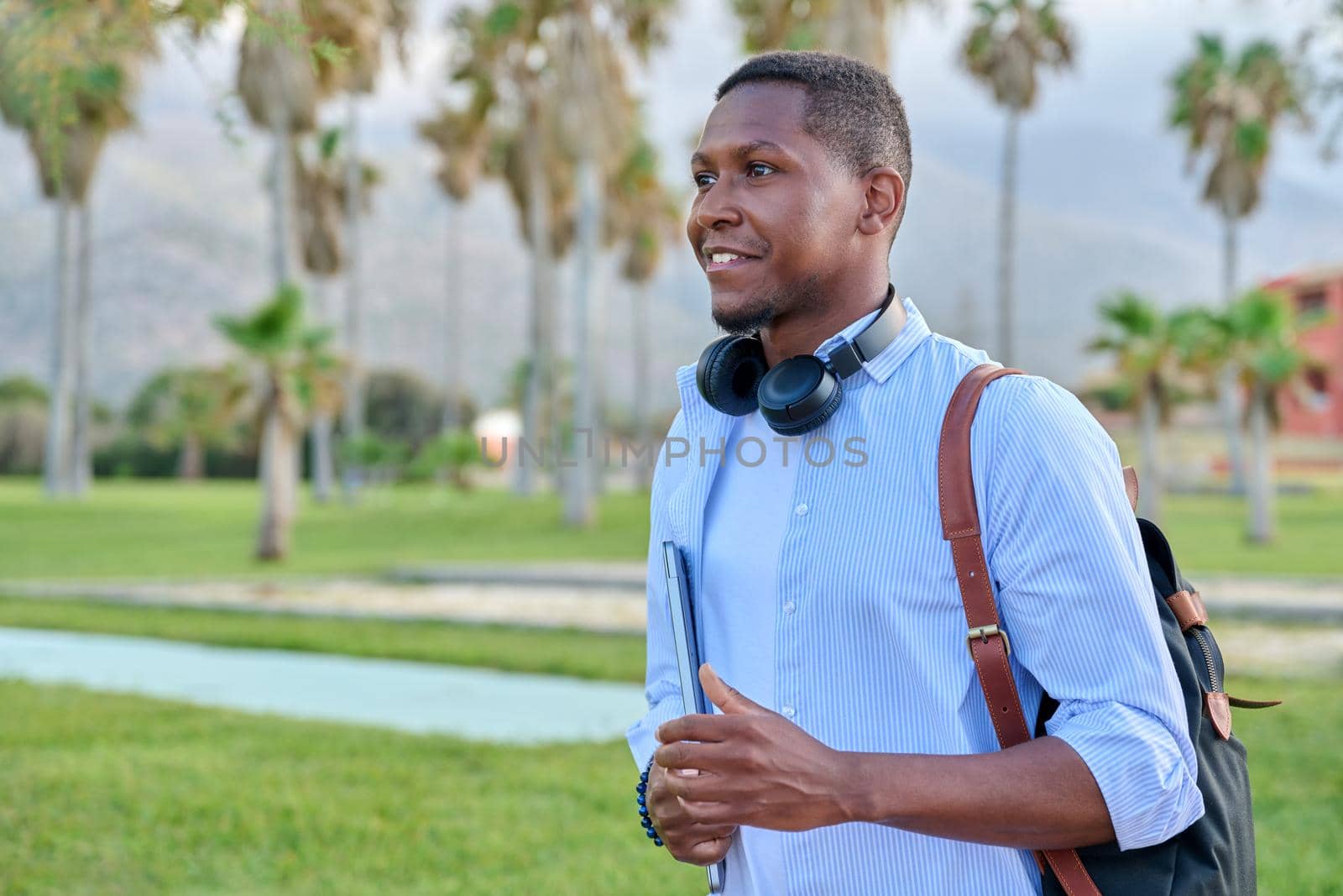 Outdoor portrait of young man with laptop headphones and backpack. African male adult student, creative freelancer, businessmen in tropical park. E-learning, online technologies, education, business