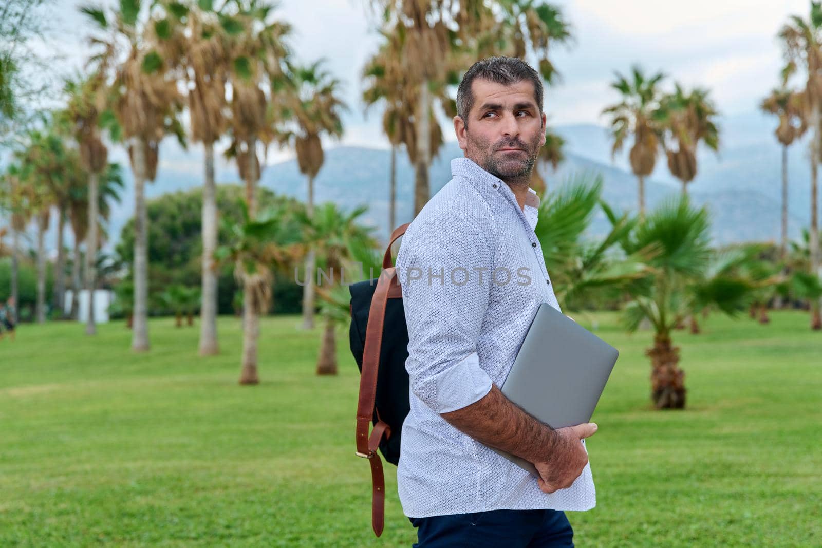 Portrait of serious confident mature male with laptop outdoors. Businessman with backpack, laptop in hands, tropical park nature. Business, technology, freelance, lifestyle, middle age, business trip