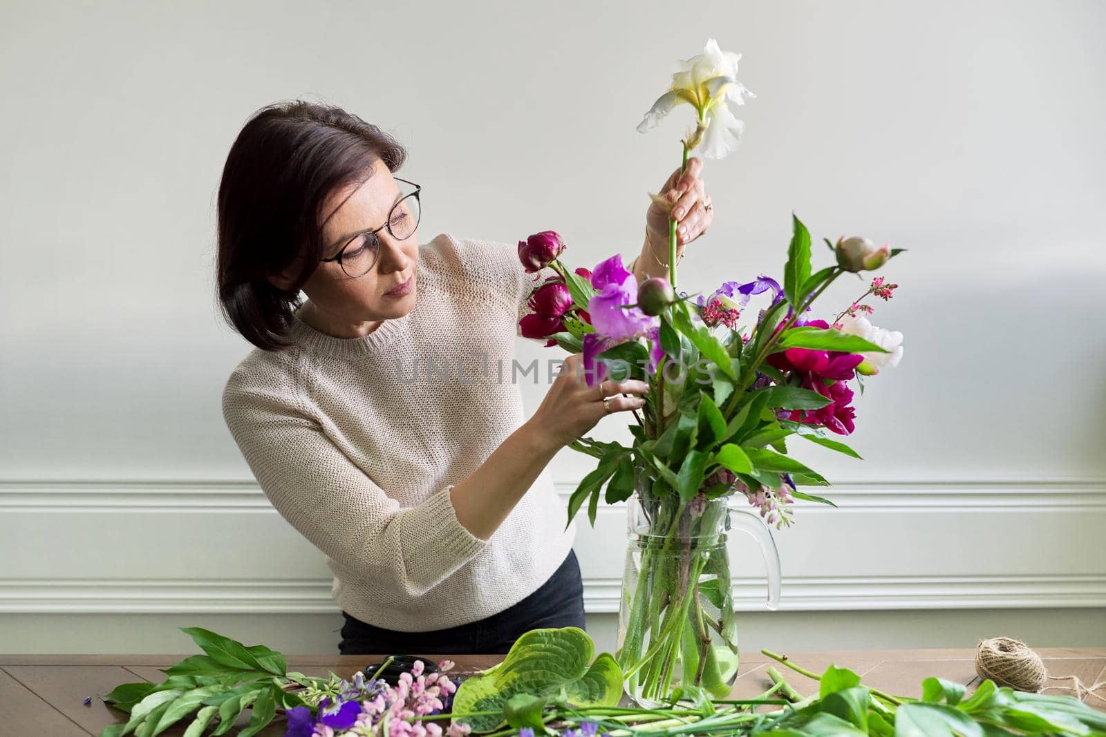 Mature woman at home with spring flowers. Female making bouquet, placing flowers in jug of water. Spring summer season, floristry, home decoration, natural beauty concept