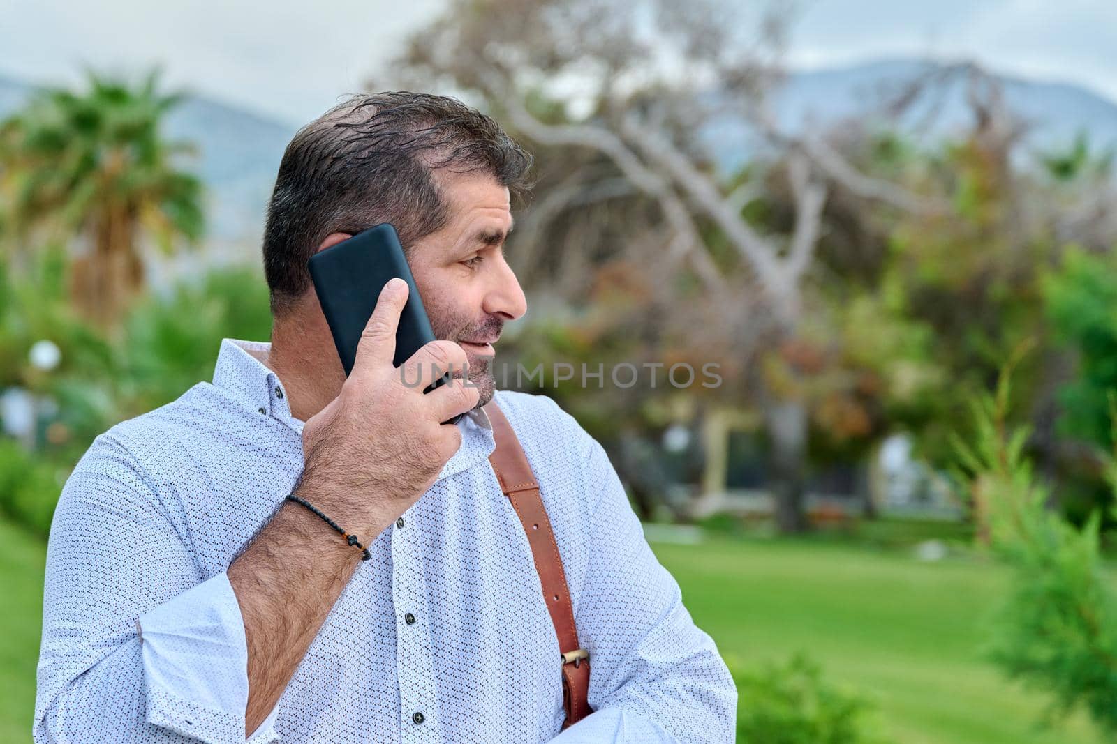 Mature business man talking on the phone outdoors. Serious confident male with backpack, in tropical park. Business trip, freelance, communication, technology, middle aged people concept, copy space