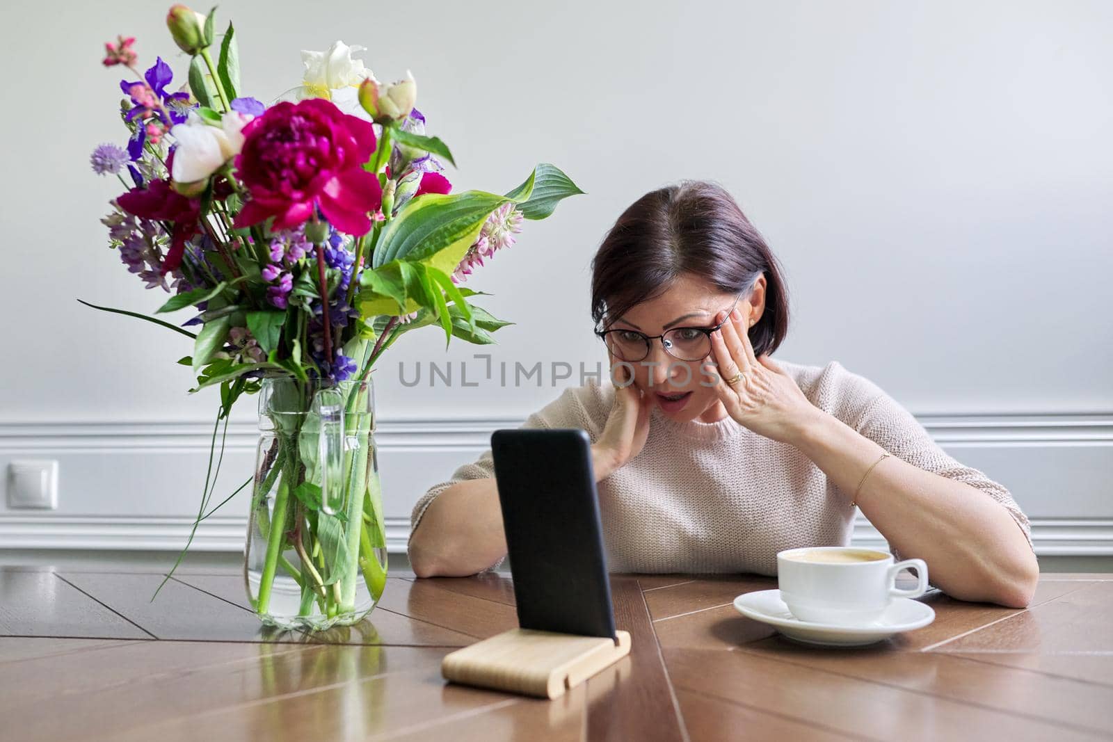 Surprised emotional mature woman looking at smartphone screen sitting at table at home