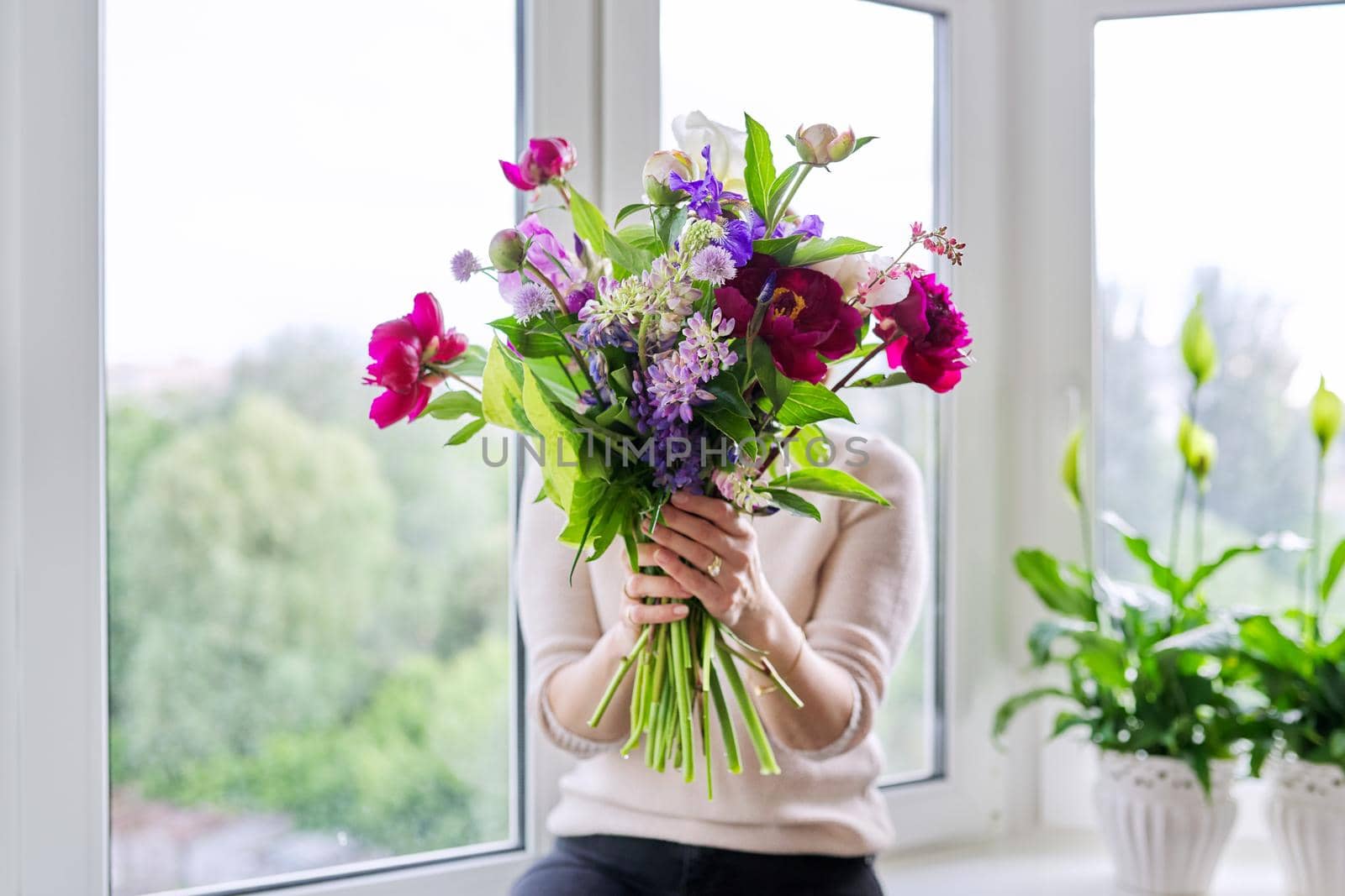 Bouquet of bright flowers of peonies irises lupins in the hands of woman close-up, female at home