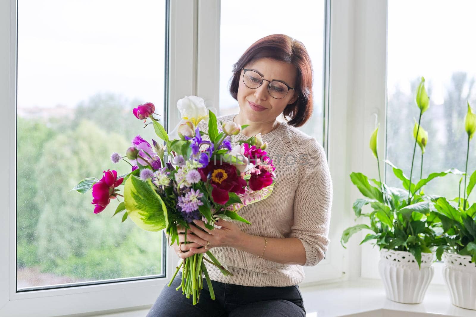 Portrait of beautiful mature woman with bouquet at home. Smiling female with flowers and buds of peony iris lupine. Spring summer season, natural beauty, middle aged people, gift, holiday concept