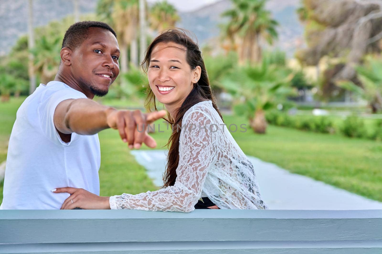 Beautiful young multi-ethnic couple sitting together on bench in tropical park. Loving asian woman and african man embracing. Male showing index finger. Relationship, happiness, multicultural family