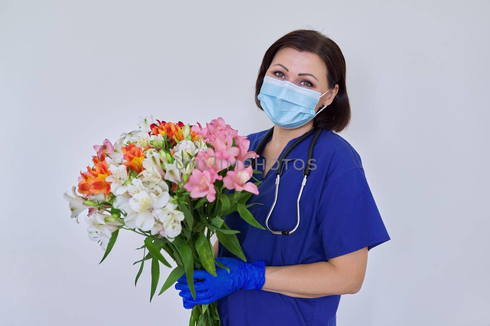 Doctor's Day, Nurse's Day. Female medic in blue uniform medical protective face mask gloves with stethoscope with large bouquet of flowers on light background