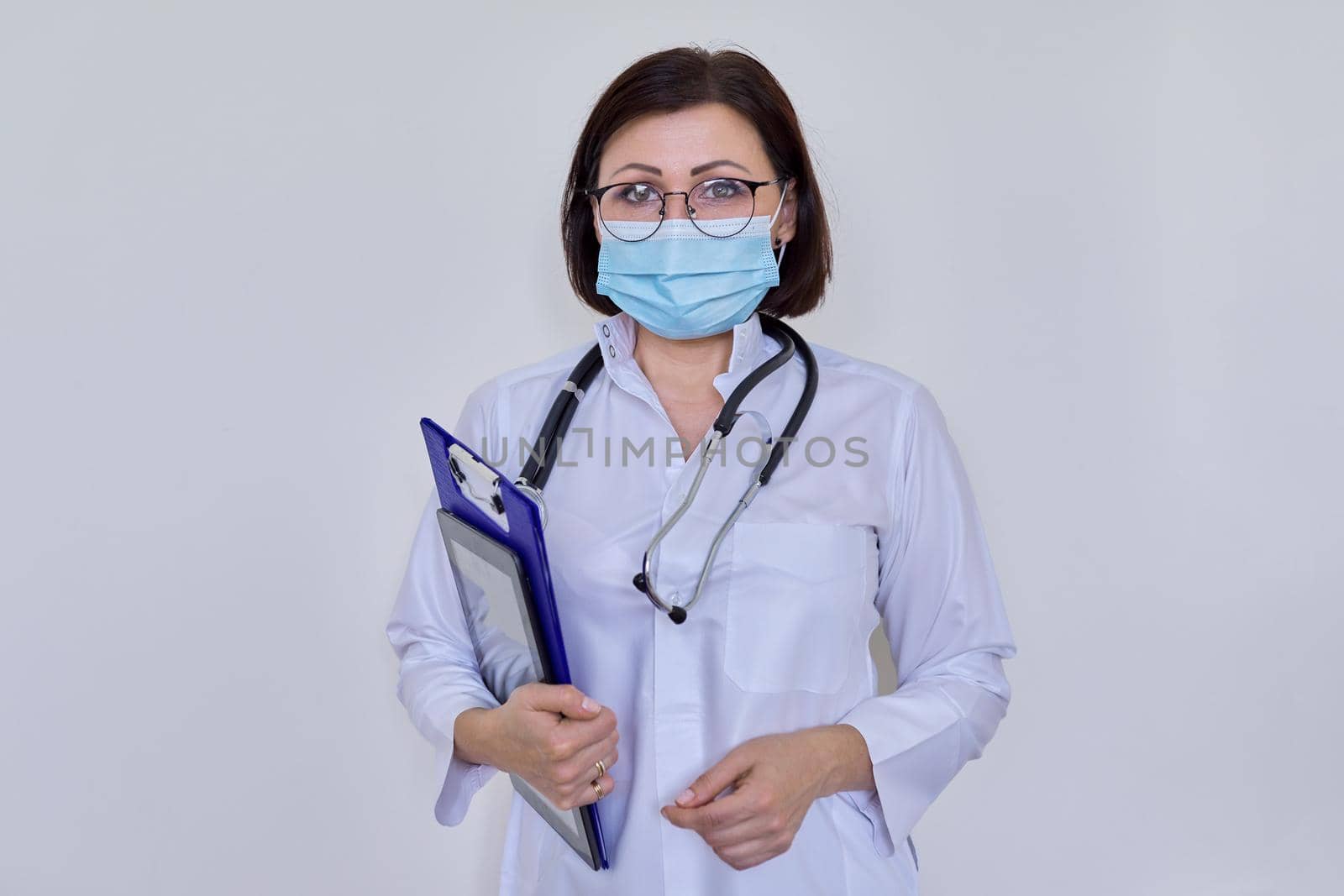 Portrait of female doctor in white uniform in protective medical face mask with stethoscope, with clipboard and digital tablet in her hands, woman looking at camera, on light background