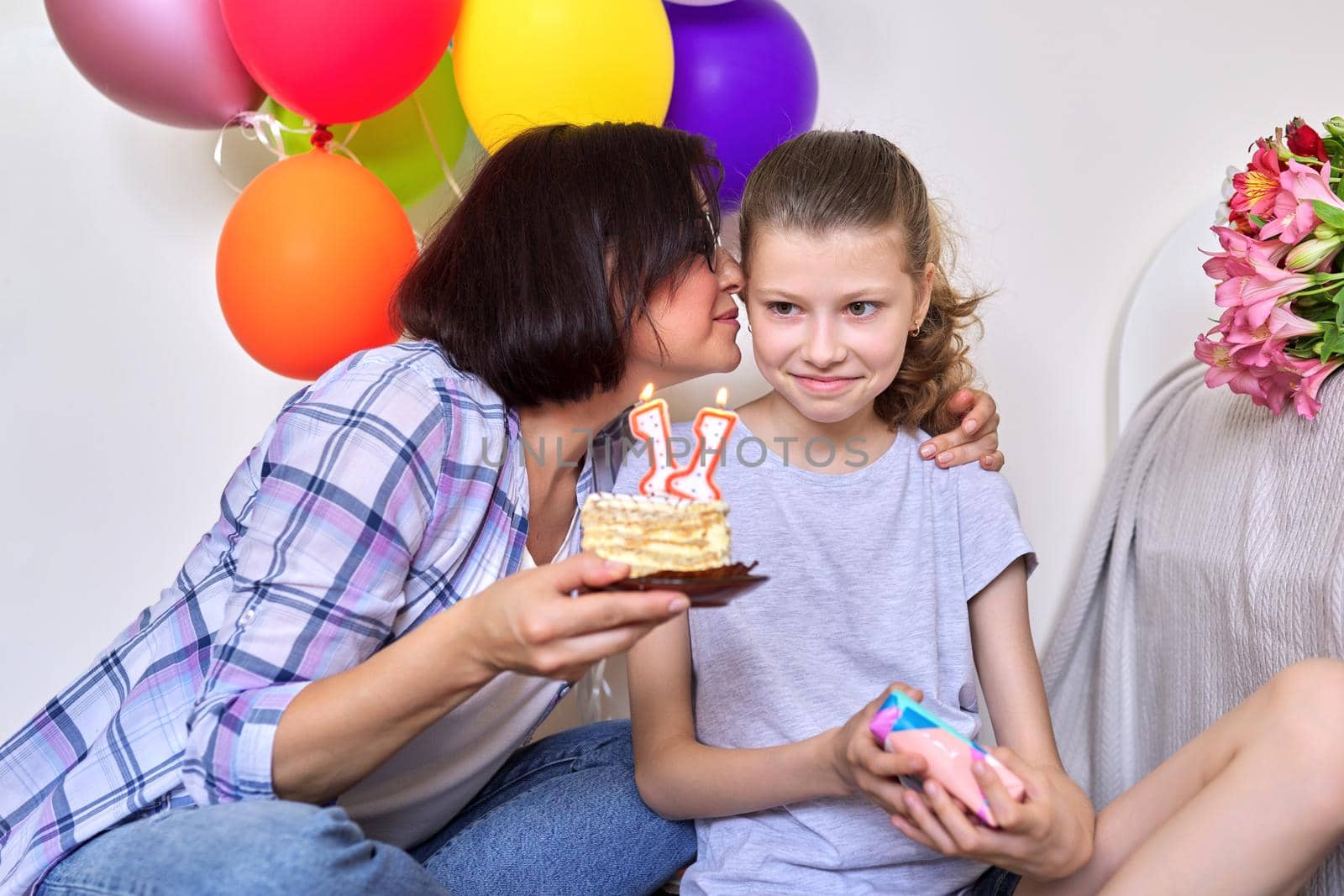 Childs birthday, 11 years old. Mom and daughter sitting together at home with small cake with candles, gift, balloons, bouquet of flowers