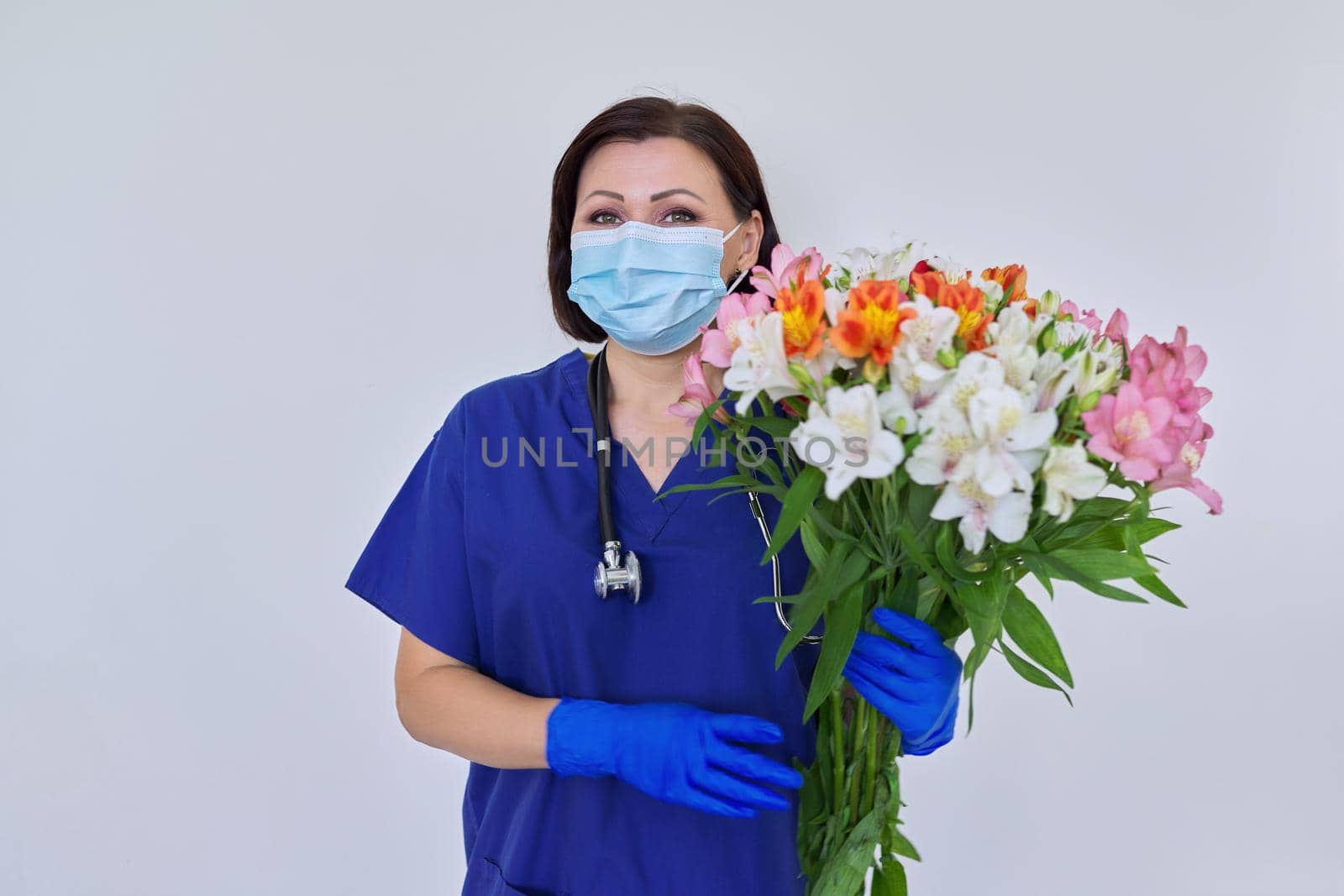 Doctor's Day, Nurse's Day. Female medic in blue uniform medical protective face mask gloves with stethoscope with large bouquet of flowers on light background