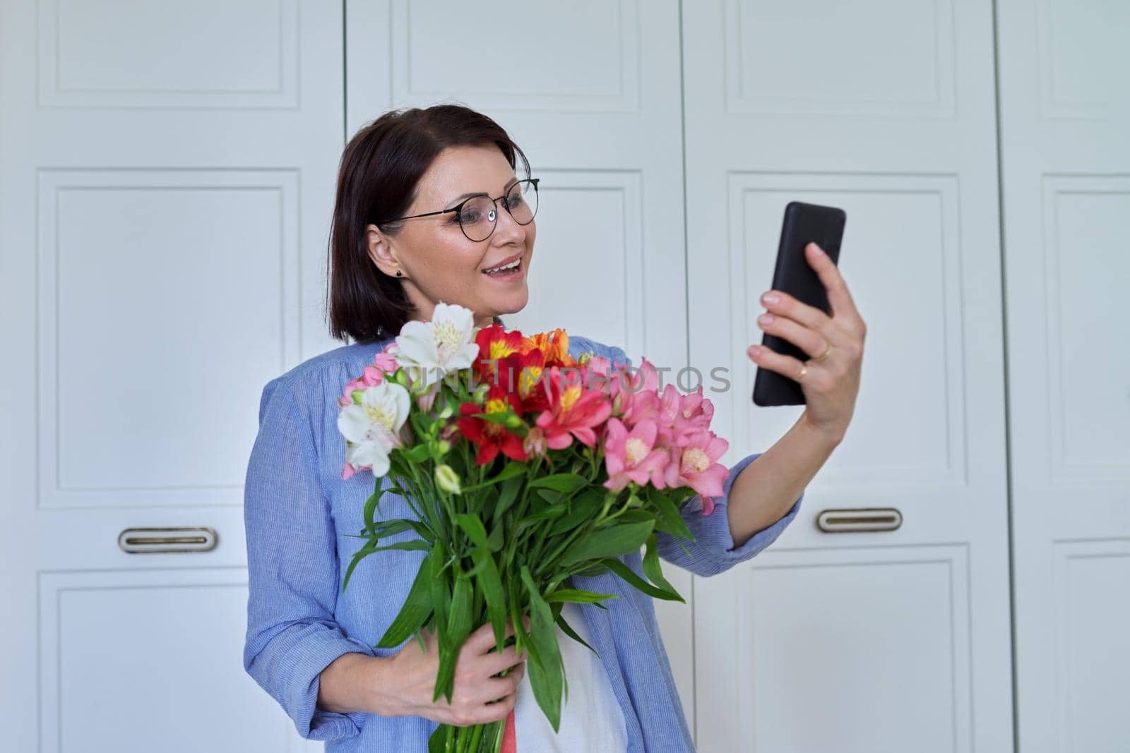 Happy middle-aged woman with bouquet of flowers and smartphone, female taking selfie, talking using video call, on light background at home. Holiday, date, birthday, happiness, joy, mature people