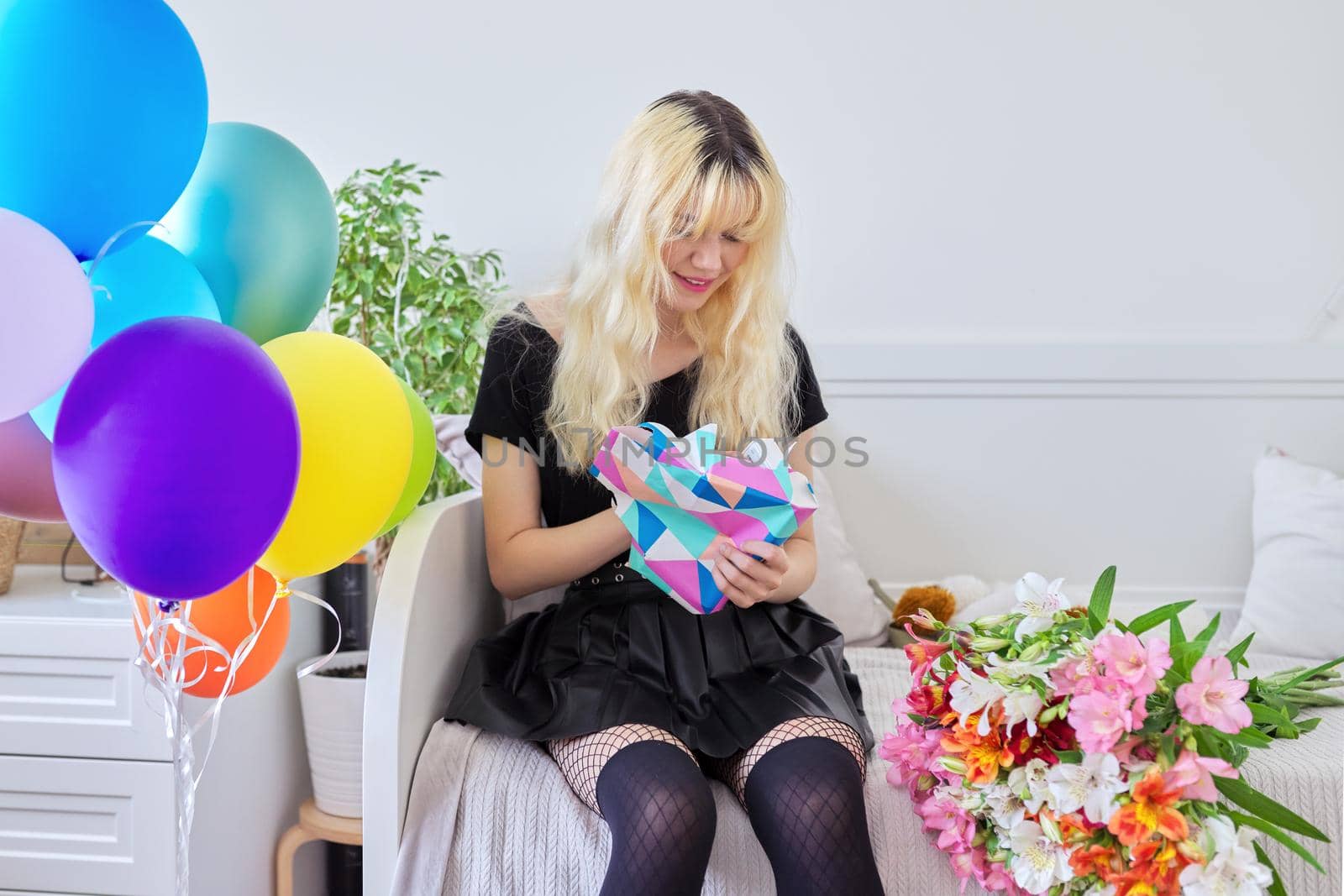 Birthday of teenage female 16, 17 years old. Hipster teenager at home with surprise gift in box, bouquet of flowers, colored balloons, sitting at home on bed. Teens, age, holiday concept