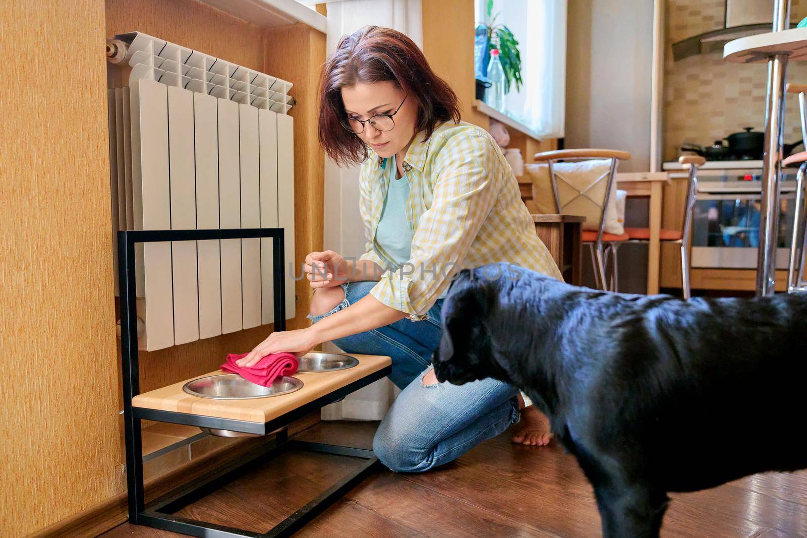 Middle aged woman and pet dog at home in kitchen interior. Pet owner cleaning near food bowls, black labrador puppy 4 months old. Lifestyle, love, pets, 40s people concept