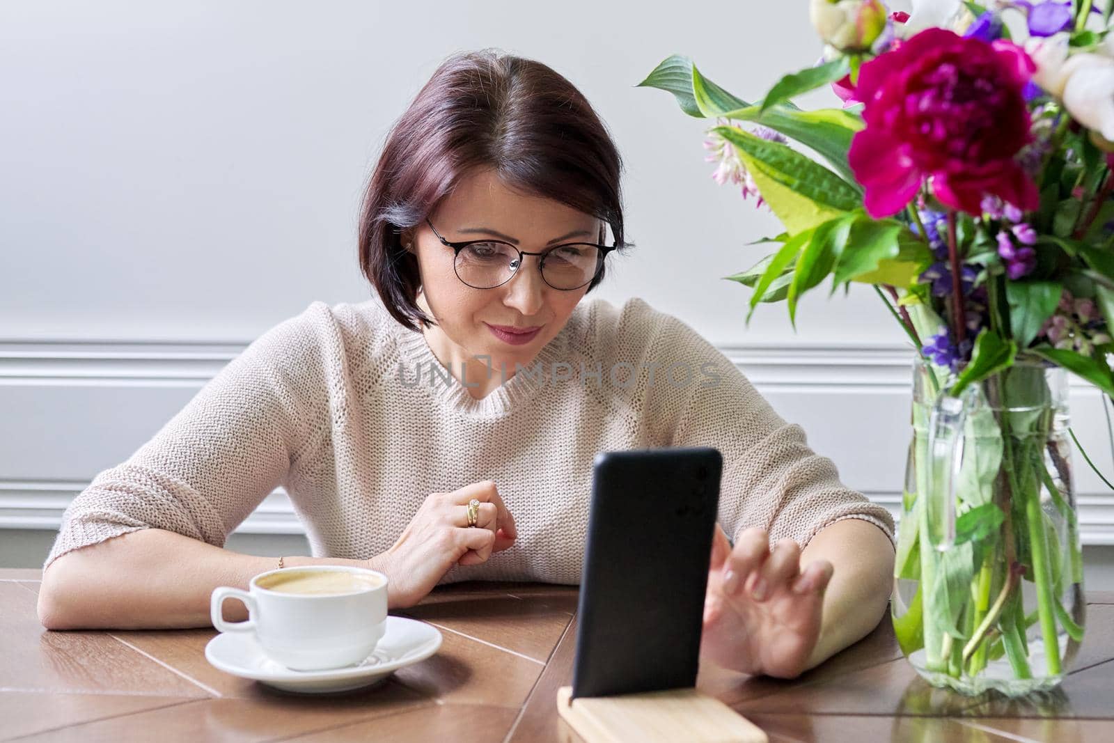 Middle-aged woman sitting at table looking at smartphone screen, female relaxing with cup of coffee, using technologies for relaxation. Lifestyle, people of mature age, home, technology concept