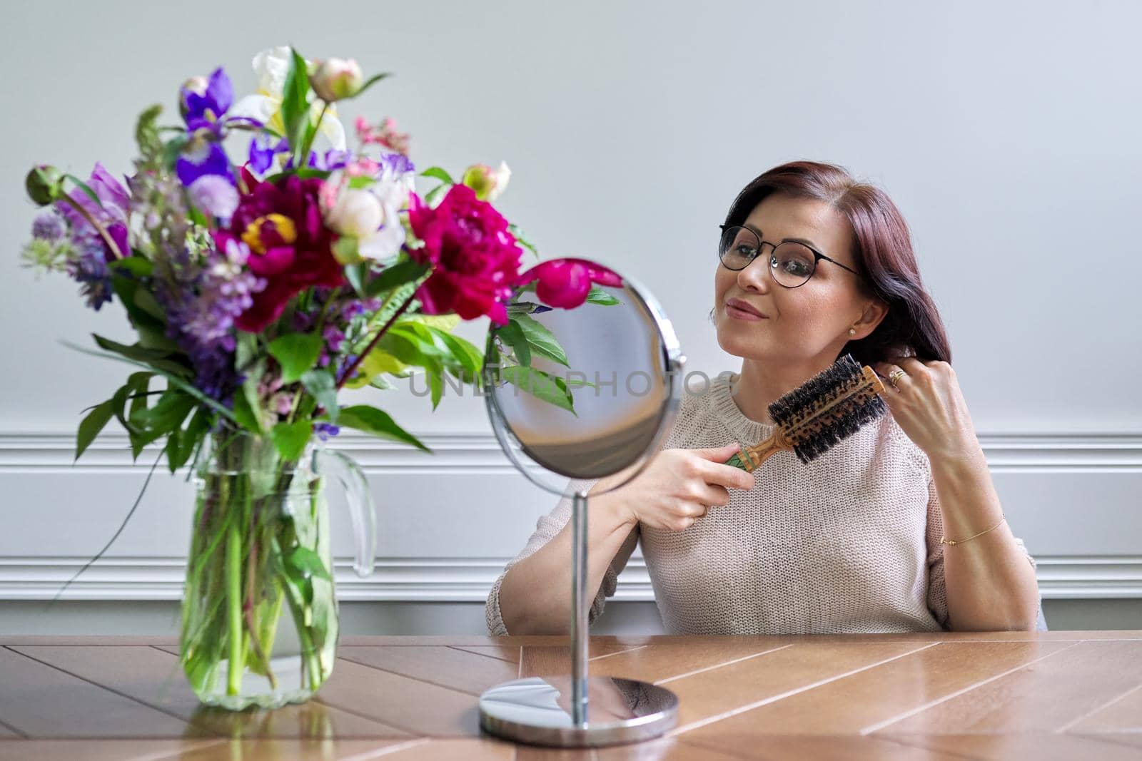 Middle aged woman styling her hair with round comb in front of makeup mirror at home by VH-studio