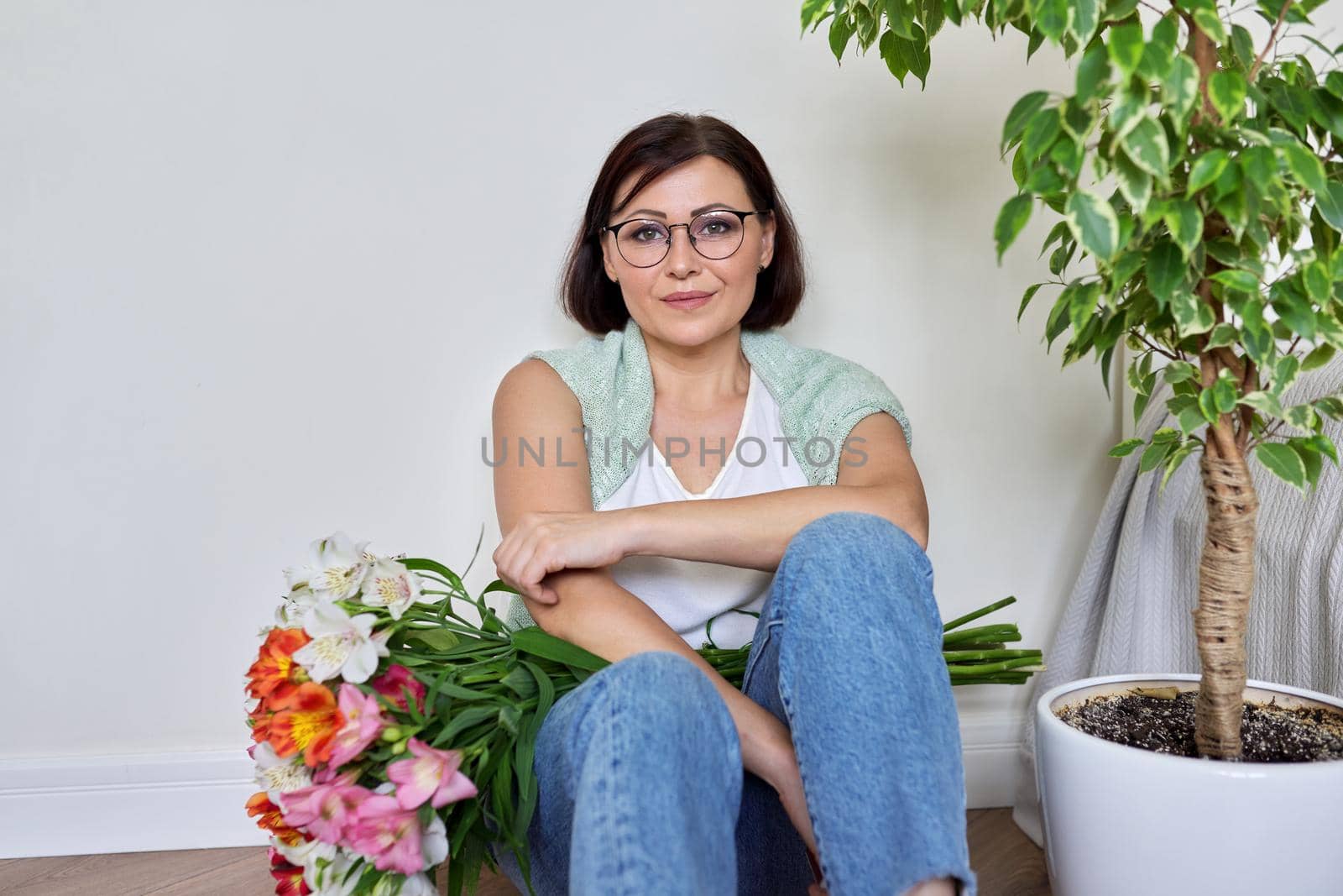 Smiling middle aged woman with bouquet of flowers sitting on the floor at home. Birthday, mother's day, holiday, mature people concept