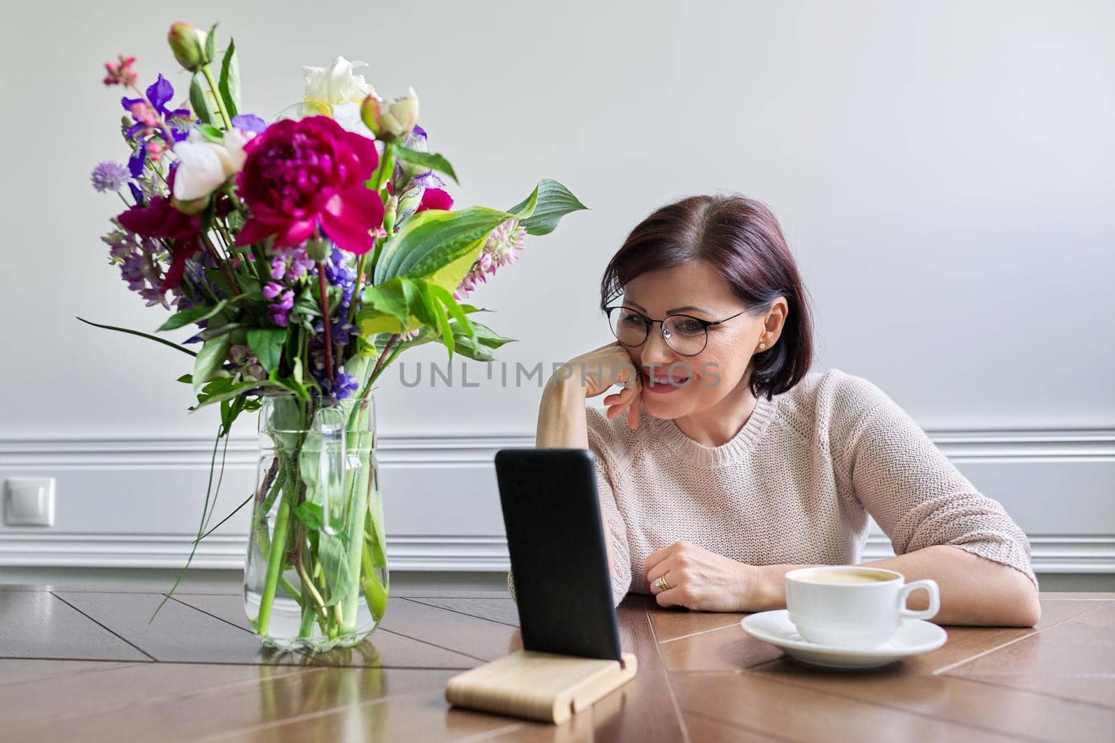 Middle-aged woman sitting at table looking at smartphone screen, female relaxing with cup of coffee, using technologies for relaxation. Lifestyle, people of mature age, home, technology concept