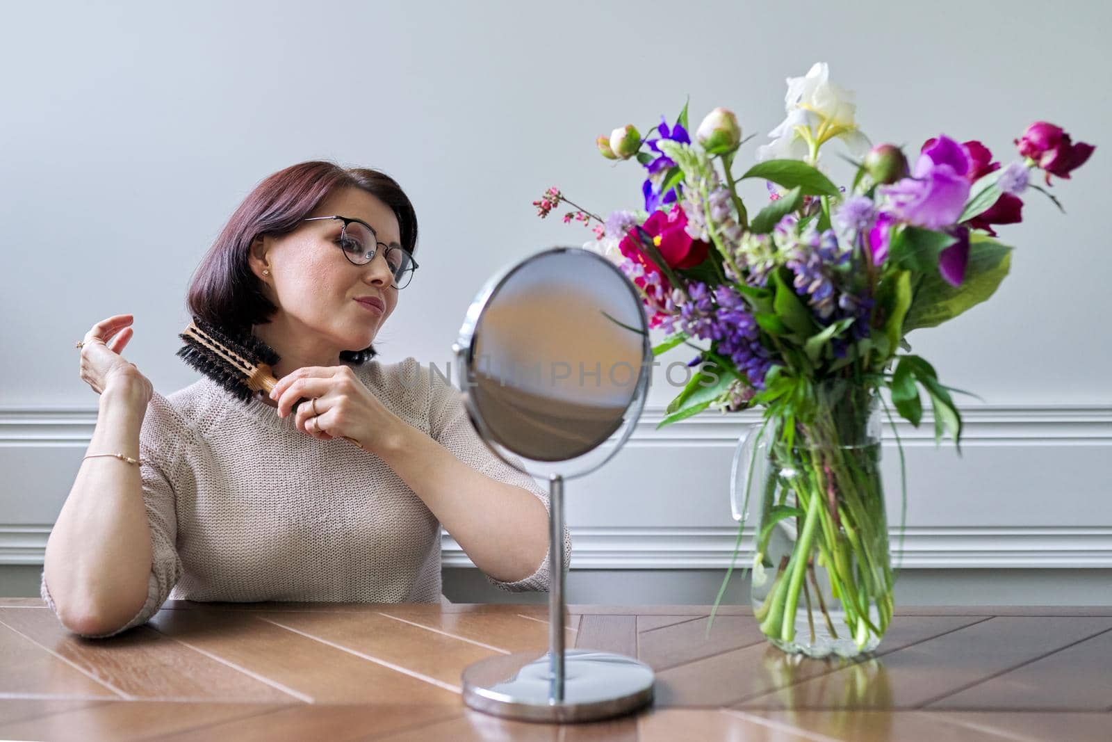 Middle aged woman styling her hair with round comb in front of makeup mirror at home at table. Beauty, hair, care, mature people concept