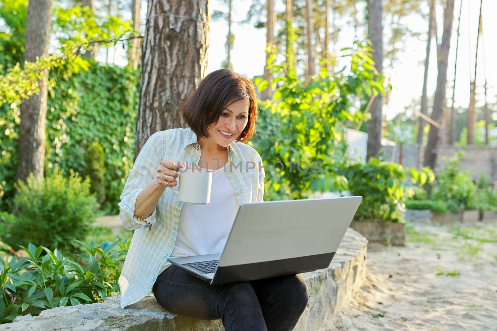 Beautiful emotional middle aged woman relaxing in garden with cup of tea and laptop. Female using video call for virtual meeting, watching movies, reading, having fun