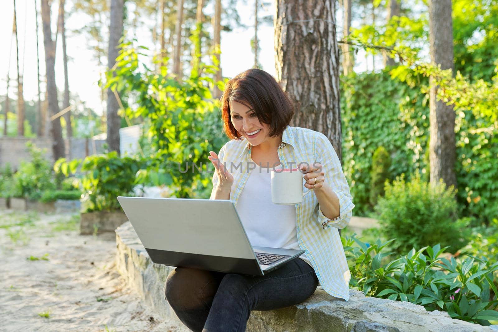 Beautiful emotional middle aged woman relaxing in garden with cup of tea and laptop by VH-studio