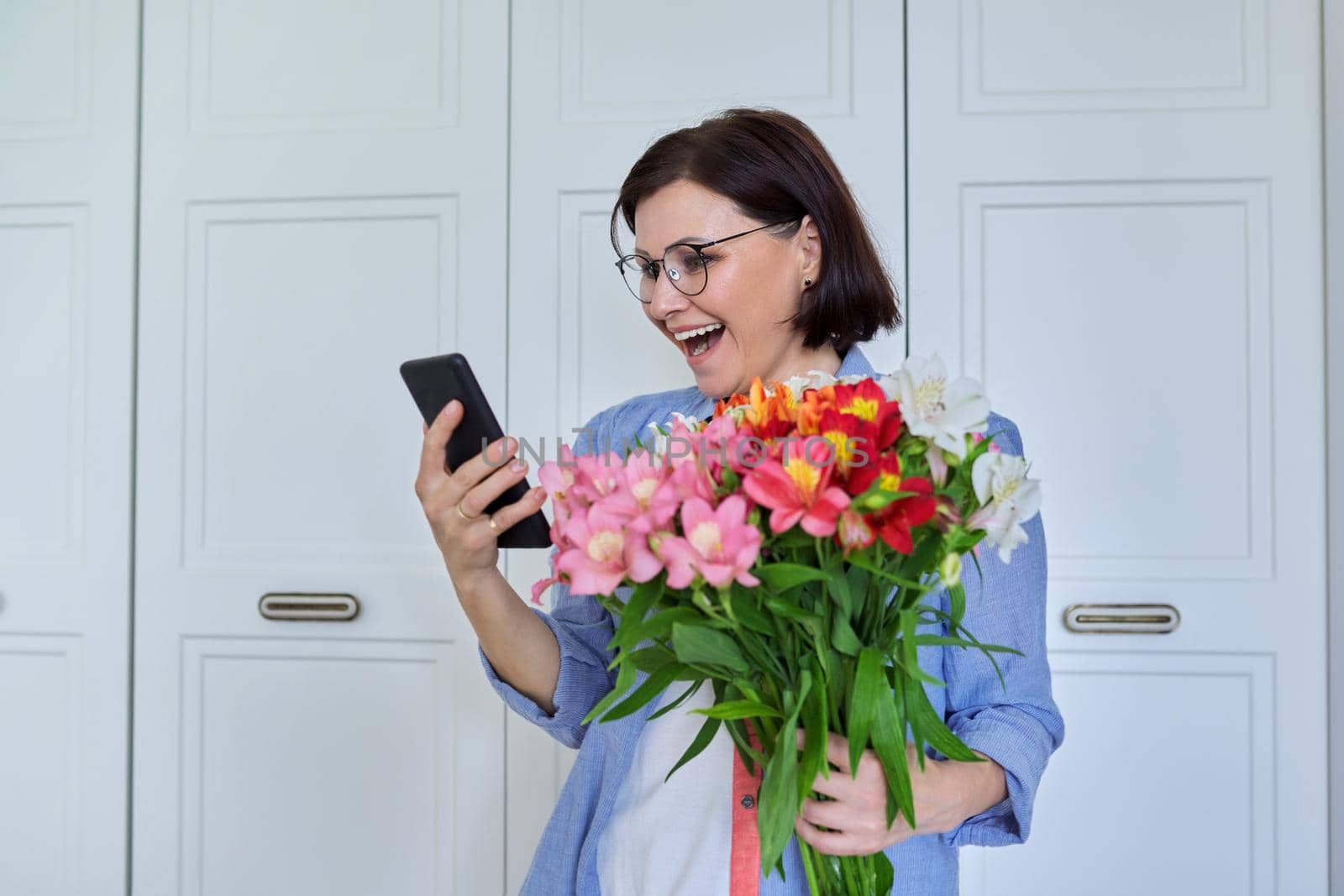 Happy middle-aged woman with large bouquet of flowers and smartphone by VH-studio