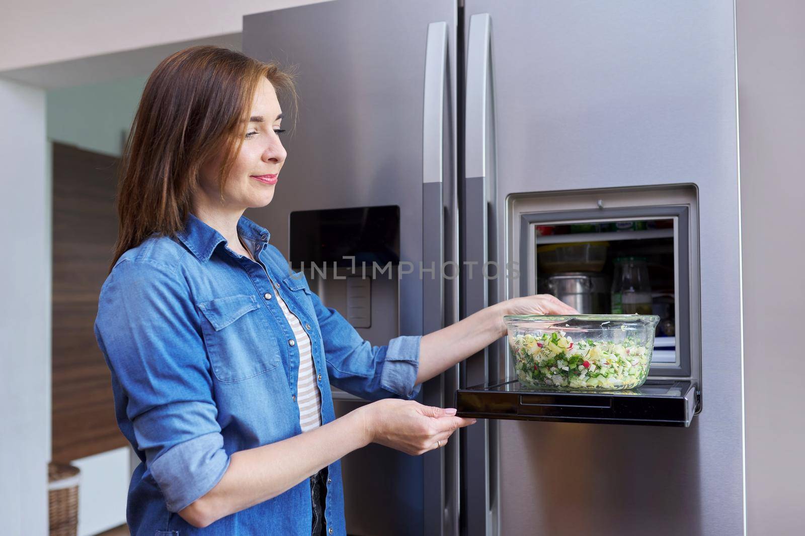 Woman with bowl of vegetable salad from refrigerator. Kitchen interior, open modern chrome refrigerator door background. Eating at home, lifestyle, household, dieting, healthly food concept