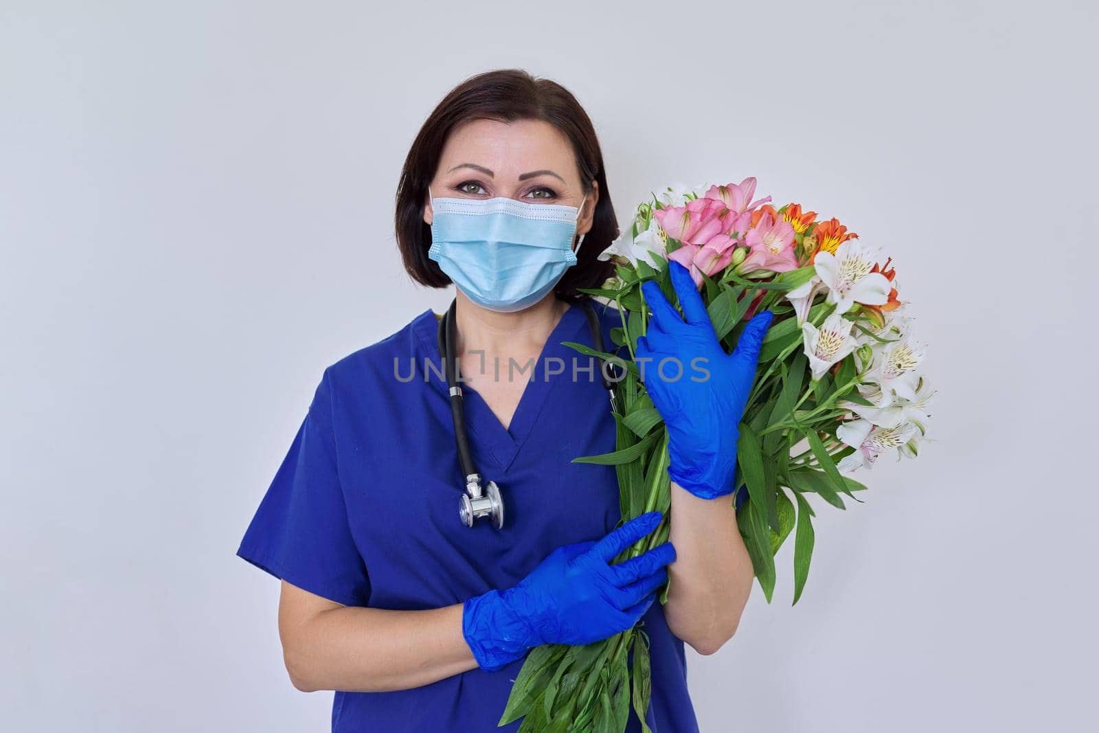 Doctor's Day, Nurse's Day. Female medic in blue uniform medical protective face mask gloves with stethoscope with large bouquet of flowers on light background