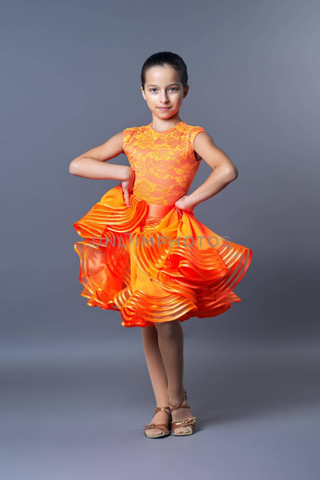 Kids sport dancing, kid girl in orange sport dress posing in dance pose looking at camera on gray studio background.